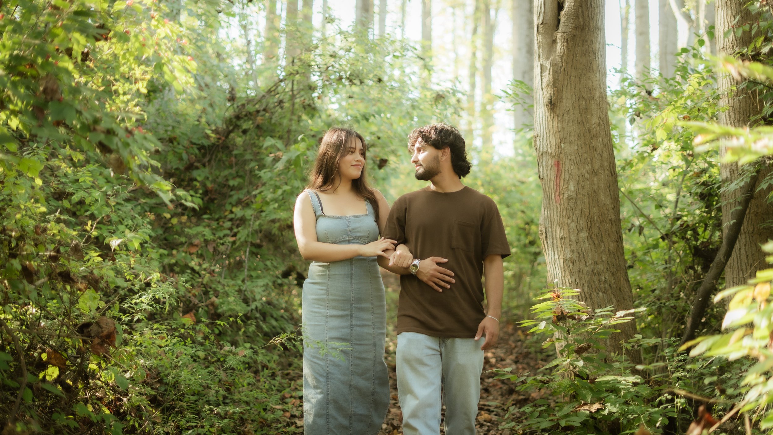 A young couple walking in a lush green forest, holding hands and looking at each other.