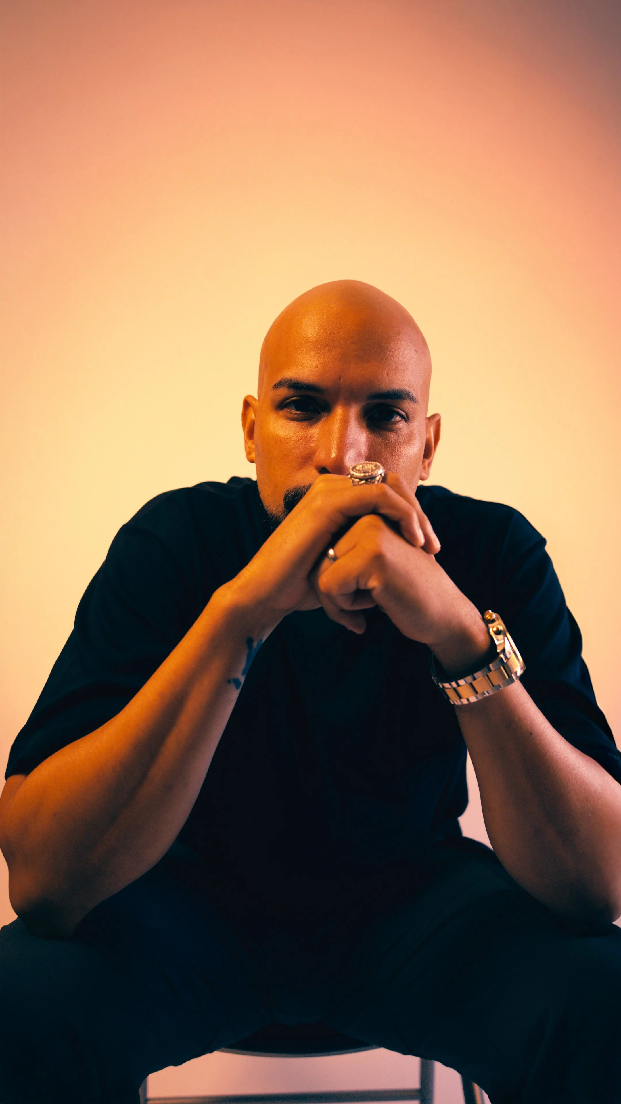 A bald man with tattoos on his arm wearing a ring, a watch, and a black shirt, sitting with his hands clasped in front of his face against a gradient background.