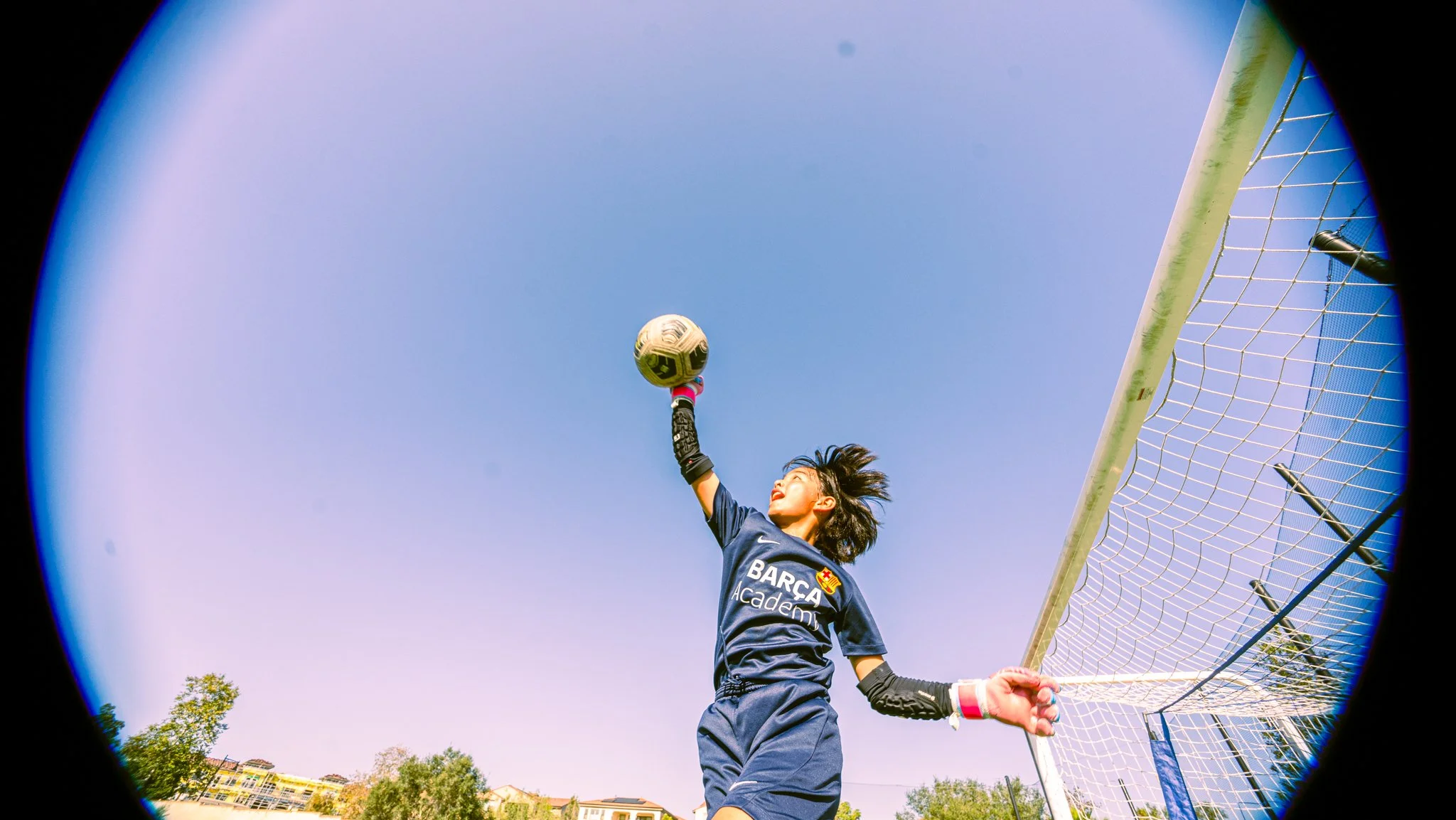 A female soccer goalkeeper jumping to catch a soccer ball near the goal post during daytime.