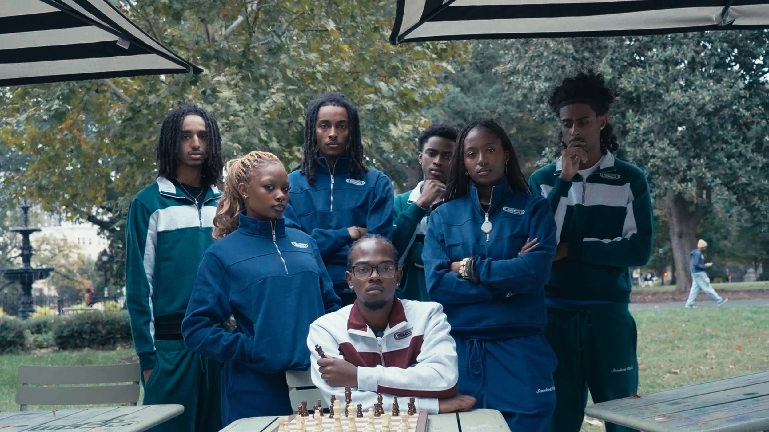 Group of seven young people, six standing and one sitting, gathered outdoors in a park, with a chessboard on a picnic table in front of them, surrounded by trees and benches.