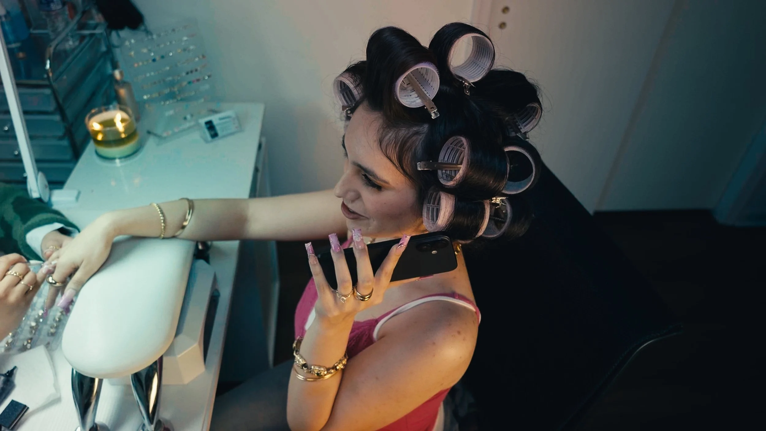 A woman with dark hair in curlers, wearing a pink tank top, speaking on a cell phone, and doing her nails at a manicure station with various nail supplies.