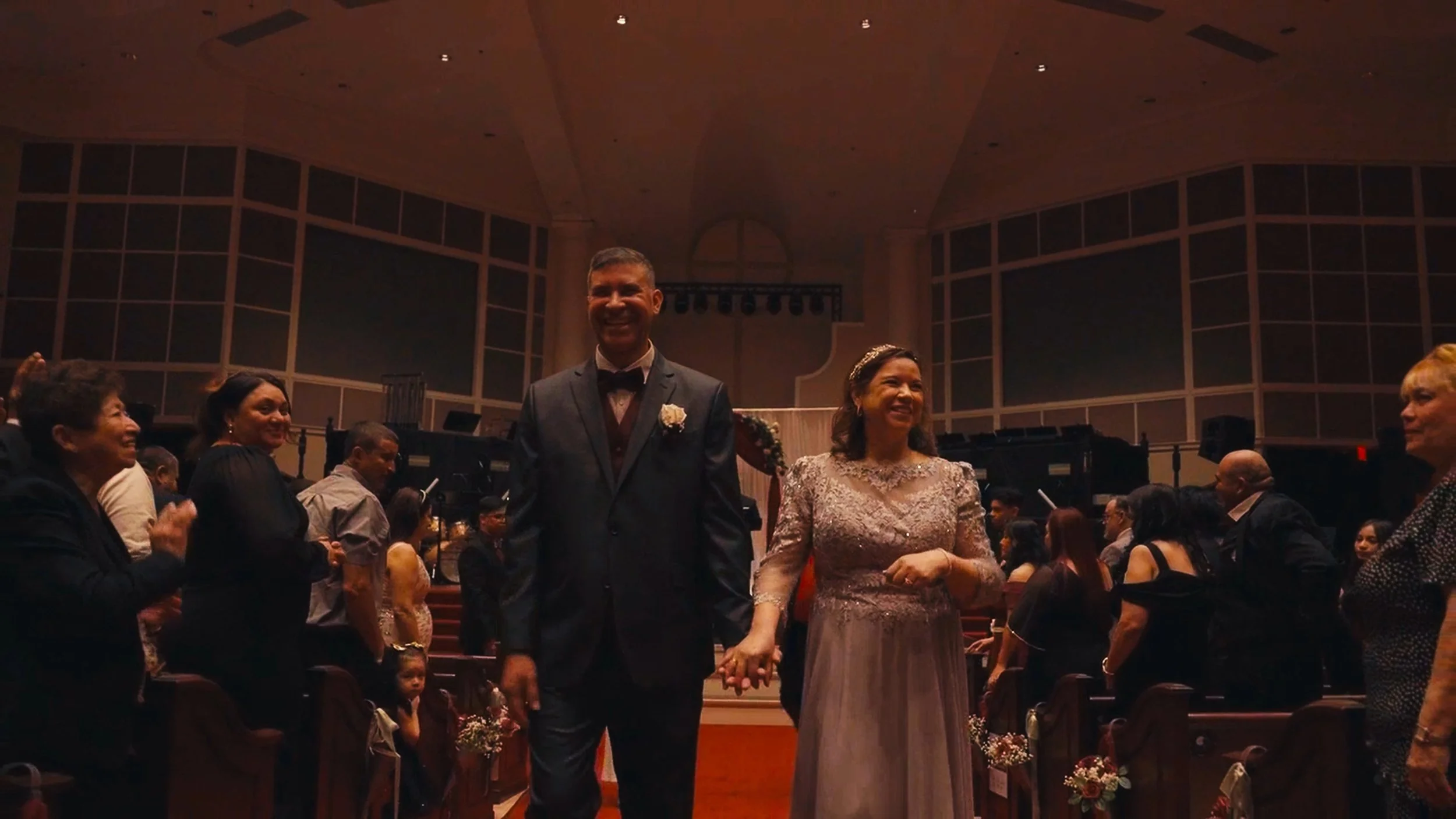 A couple dressed in wedding attire, holding hands and smiling, walking through a church or concert hall filled with seated guests, during a wedding ceremony.