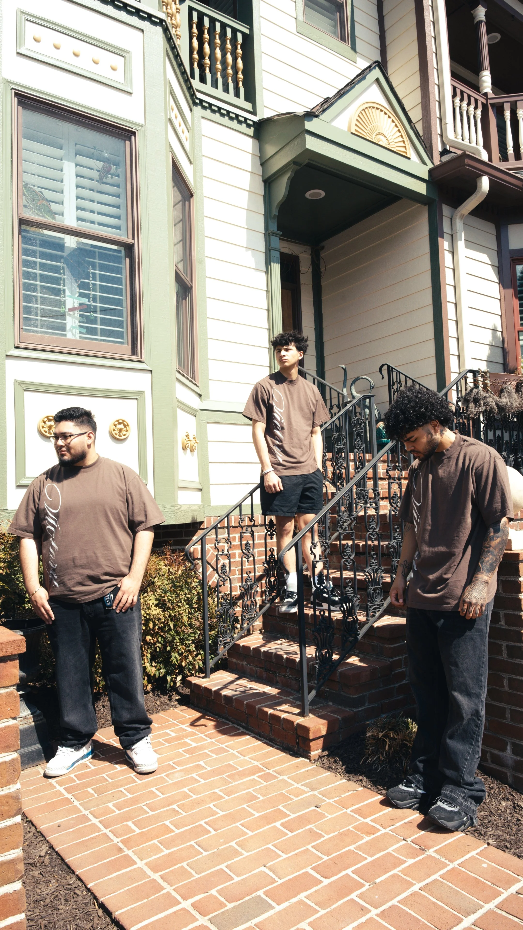 Three young men standing outside a multi-story house with a brick staircase, black iron railings, and green and beige exterior paint. They are wearing brown T-shirts and dark pants, with one looking away and others looking down or to the side in casu
