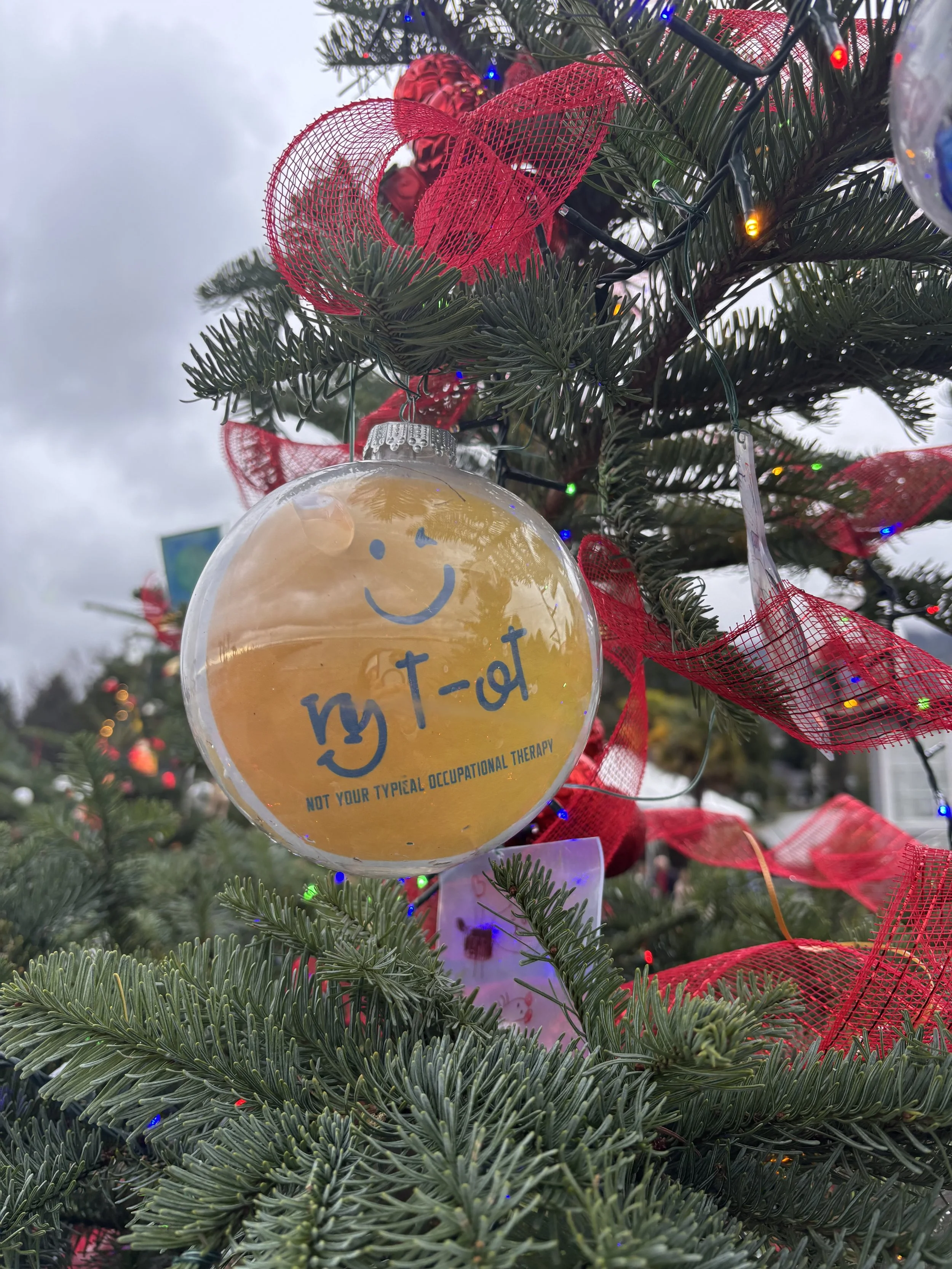 Close-up of a decorated Christmas tree with red ribbons, multicolored string lights, and a clear ornament with a yellow smiley face and the words 'not your typical occupational therapy' written on it.