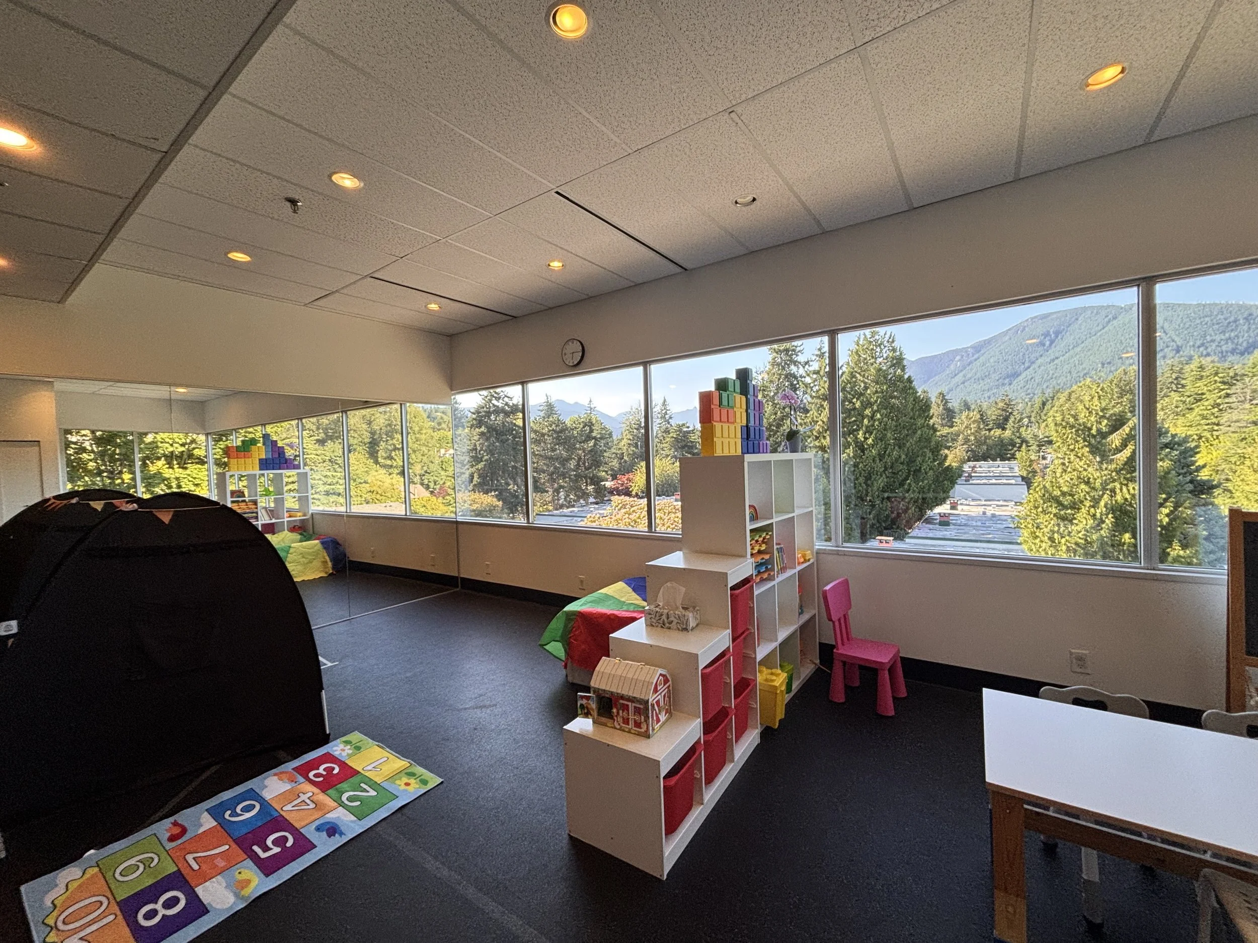 Indoor children's playroom with large windows showing trees and mountains outside. Contains colorful toys, educational mats, and small furniture.