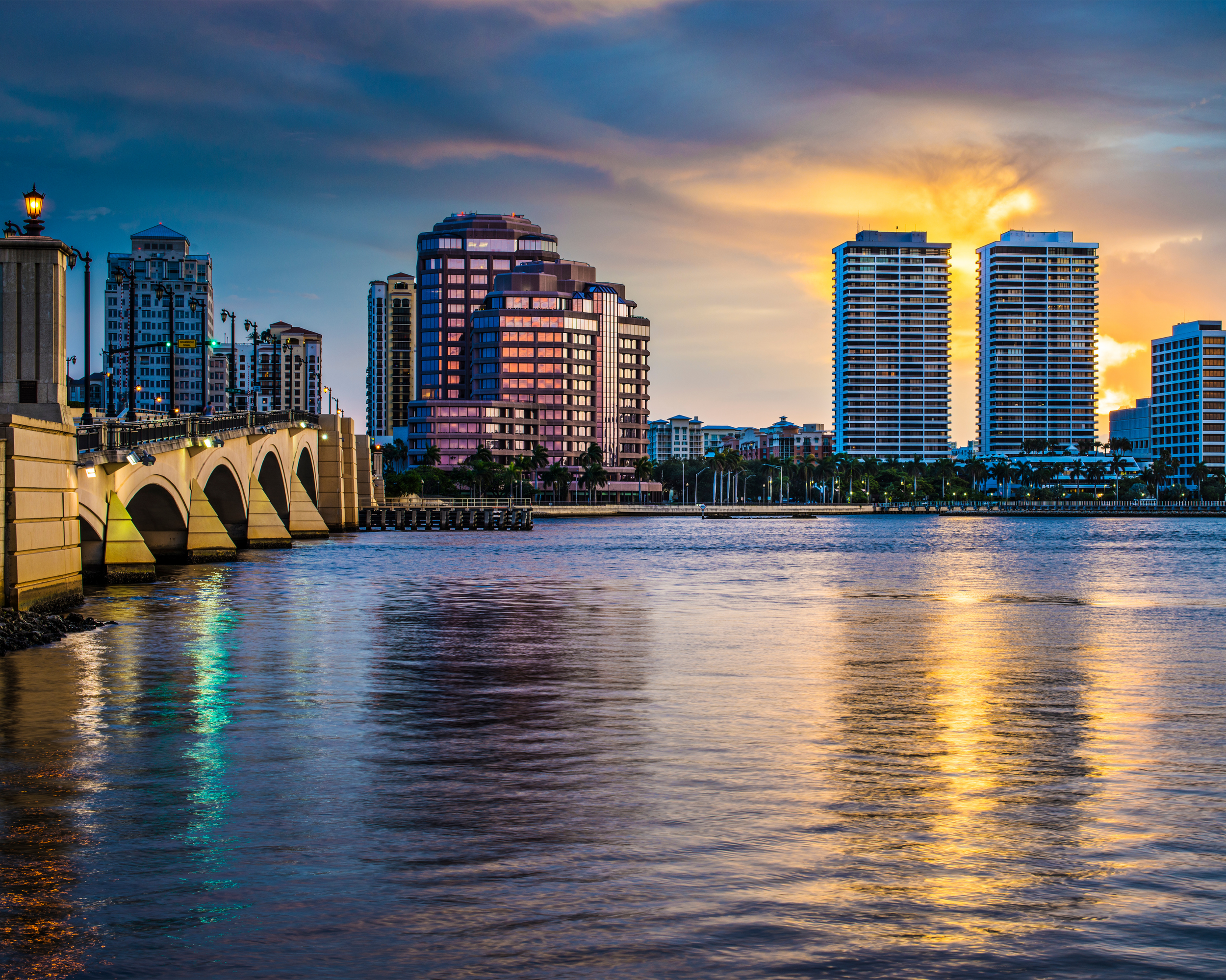 Skyline of a modern city at sunset with high-rise buildings and water reflecting the colorful sky.