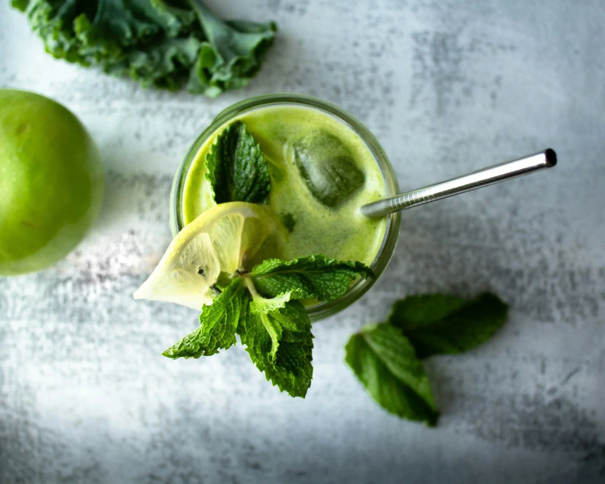 Overhead view of a glass of green smoothie with mint leaves, lemon slice, and ice cubes, surrounded by fresh lemon and mint leaves on a gray textured surface.