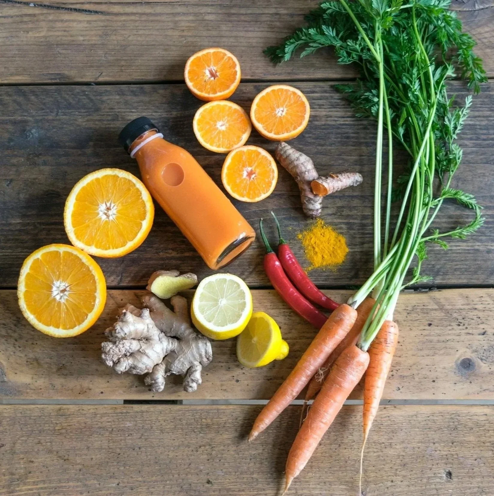Arrangement of fresh orange slices, lemon halves, ginger root, red chili peppers, carrots with green tops, turmeric powder, a small bottle of orange juice, and a piece of turmeric root on a wooden surface.