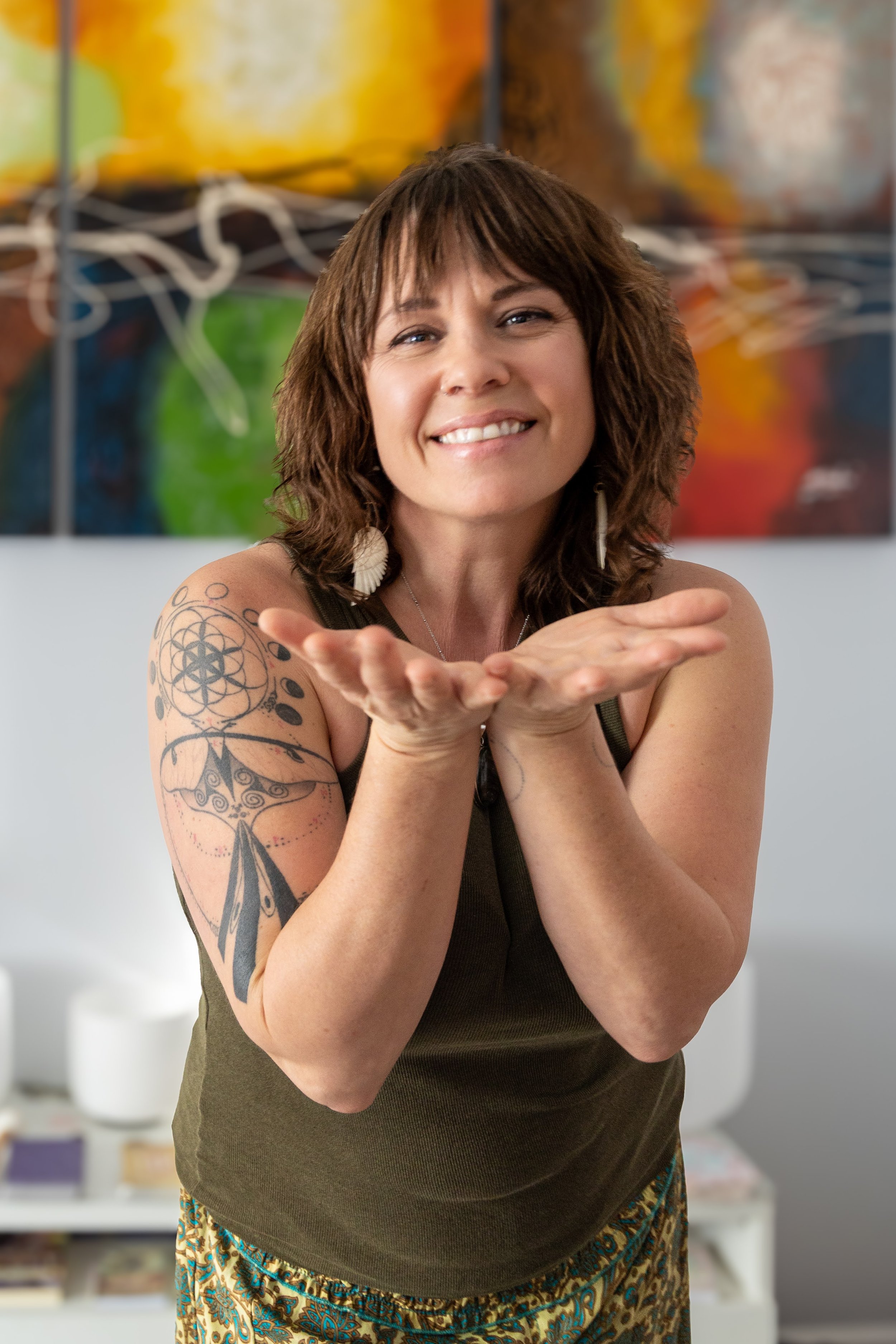 woman with tattoos smiling and holding her hands in a cupped position in front of her face, colorful abstract artwork in background