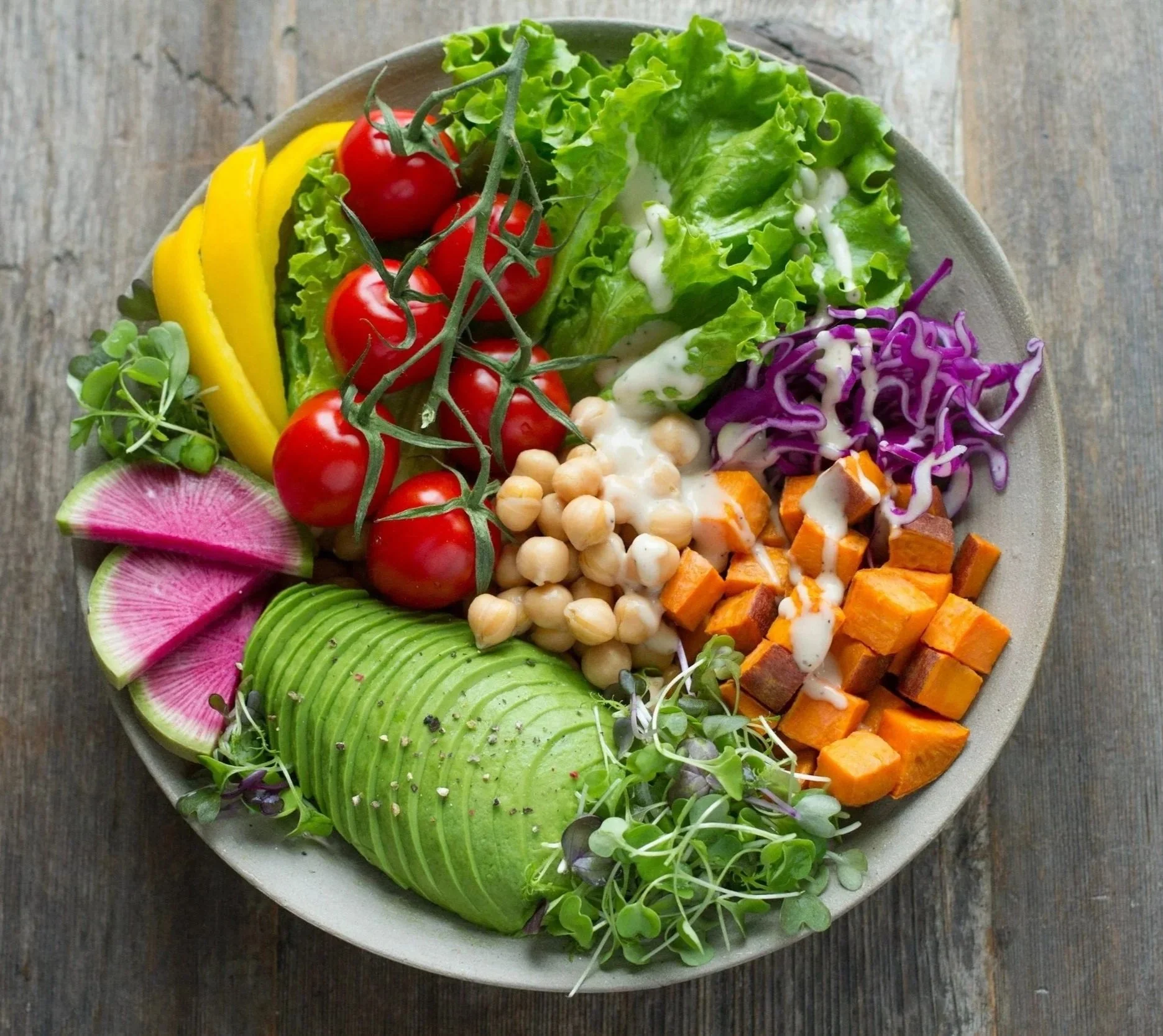 Assorted fresh vegetables in a bowl, including cherry tomatoes, lettuce, sliced radish, sliced avocado, cubed roasted sweet potatoes, purple cabbage, chickpeas, sliced yellow bell peppers, and microgreens on a wooden table.
