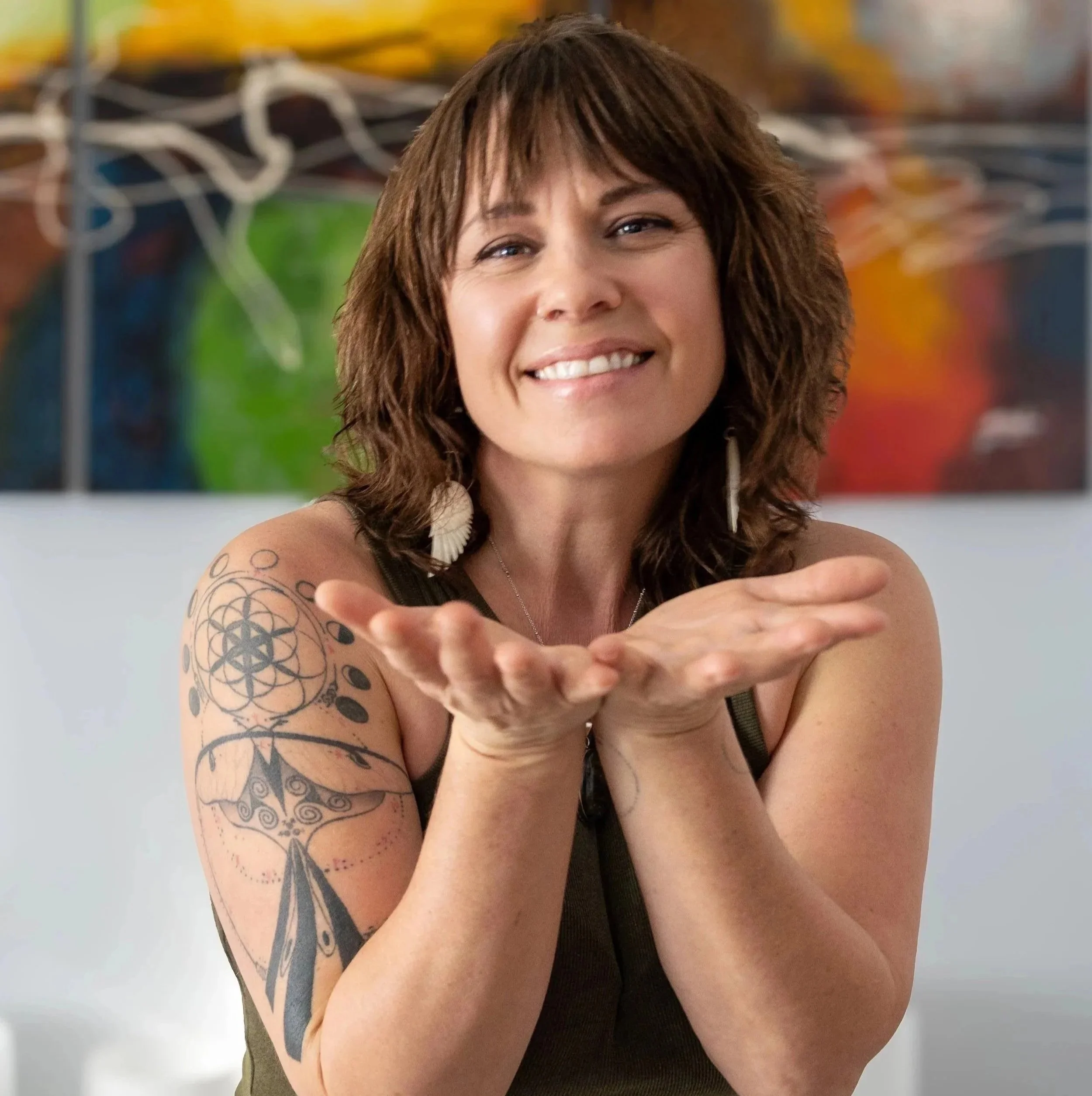 A woman with brown, wavy hair smiling and holding her hands together in front of her. She has tattoos on her upper arm and is wearing feather earrings.