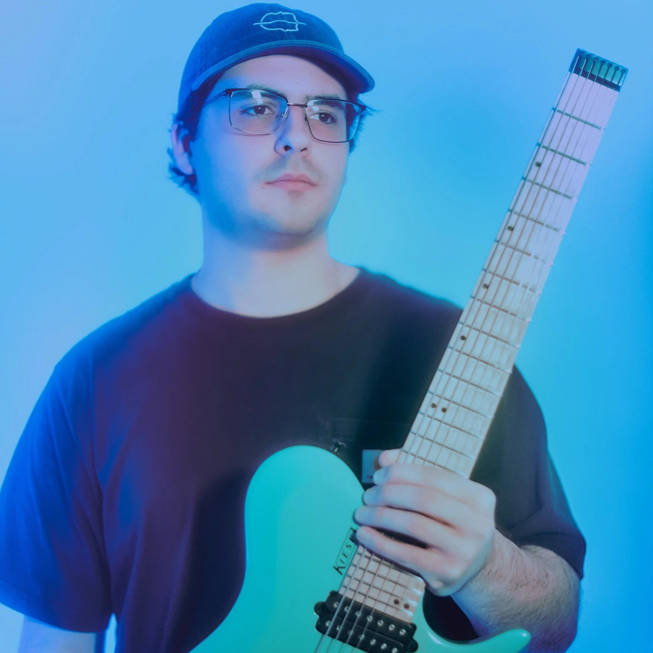 A young man wearing glasses and a baseball cap is holding an electric guitar against a blue background.