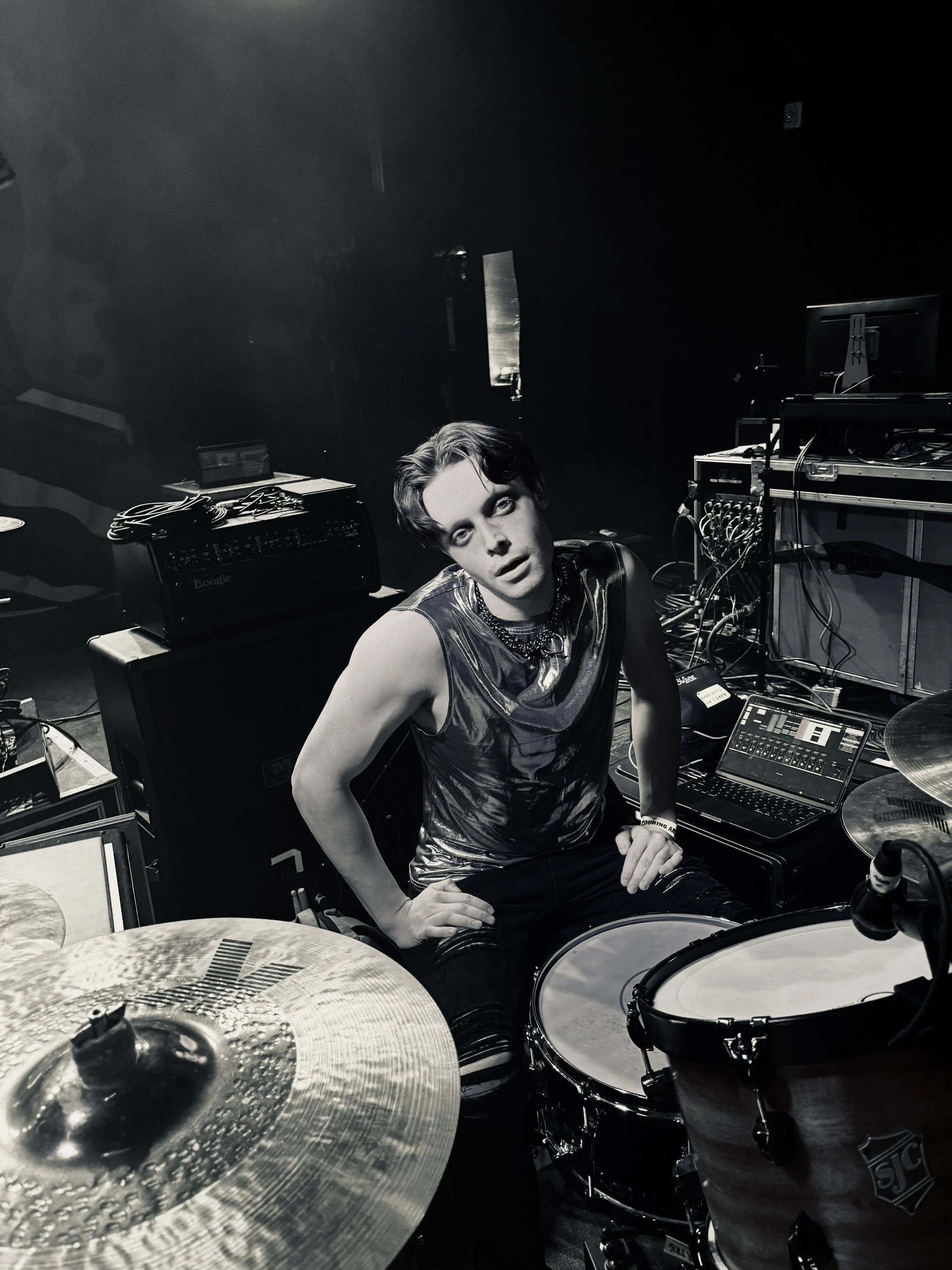 Picture of Greg Kerrigan, a content creator and touring drummer. Black and white photo of a young man sitting at a drummer's setup in a dark room, with drums, cymbals, and electronic music equipment around him.