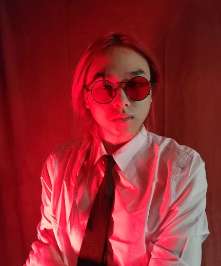 Picture of Vincent Nguyen, composer at The Espressos. Person wearing round sunglasses, white shirt, and black tie, illuminated by red lighting.