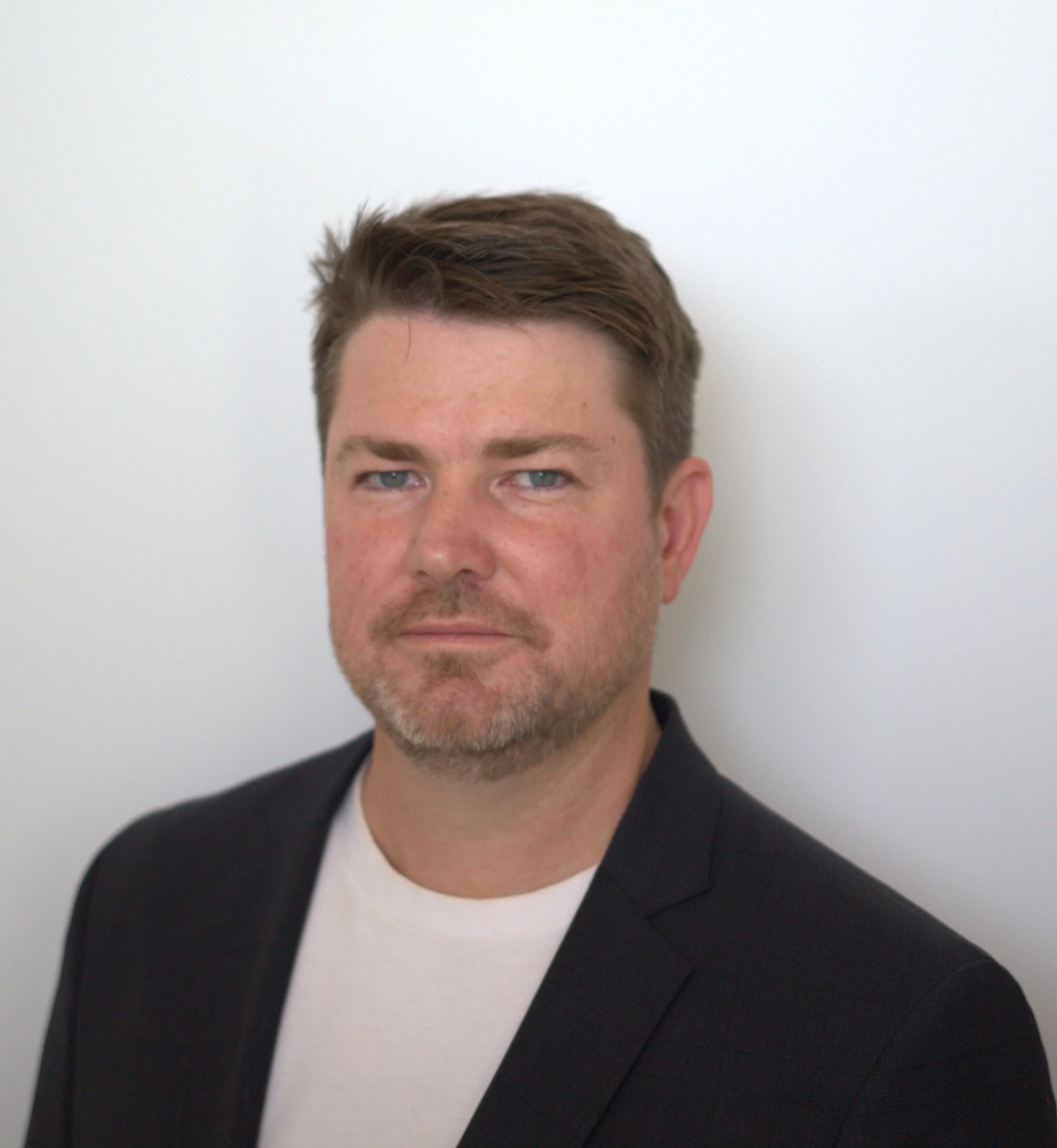 Headshot of a man with brown hair, blue eyes, and light facial hair, wearing a black blazer and white shirt, standing against a plain white background.