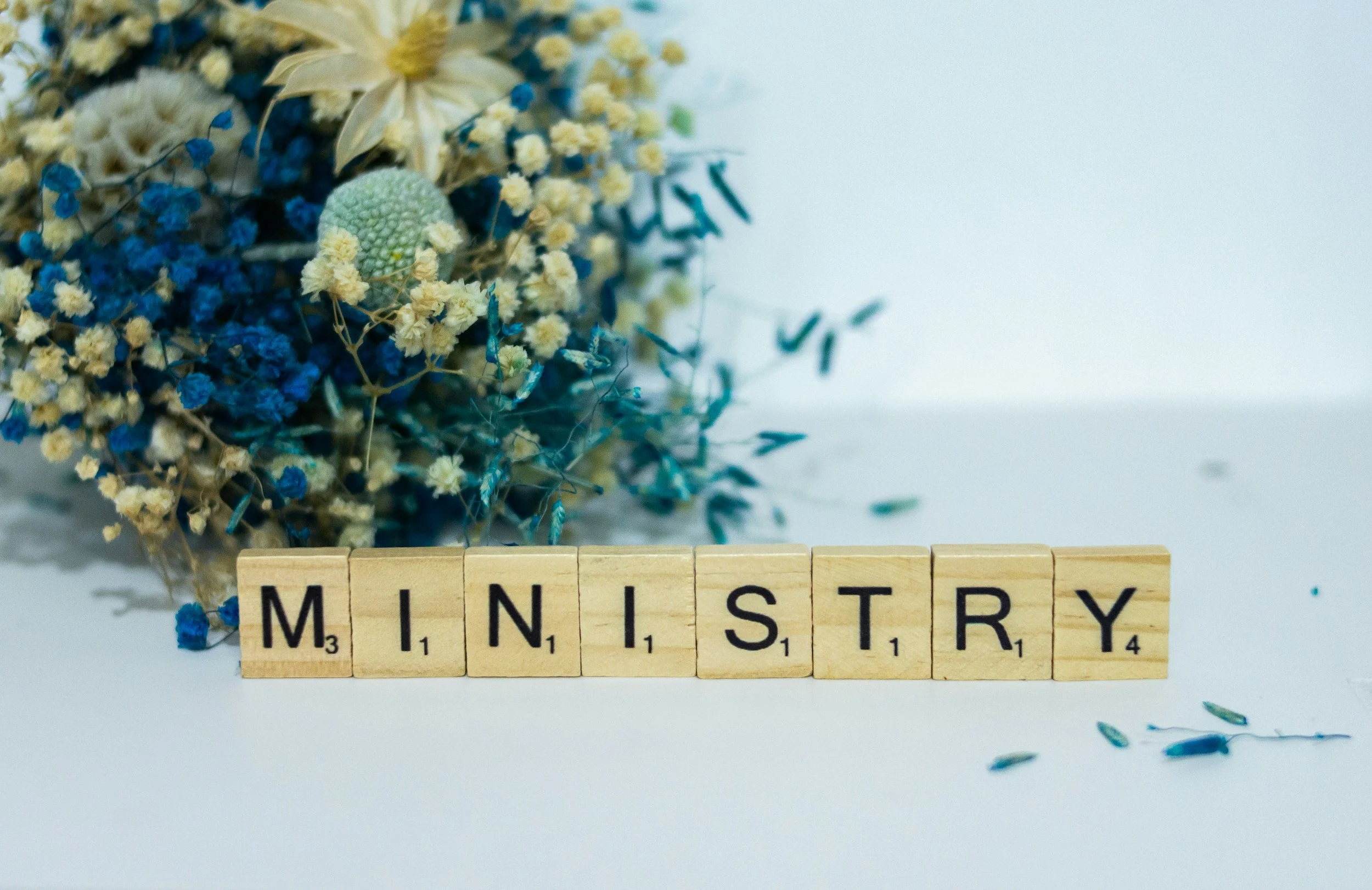 Scrabble tiles spelling 'MINISTRY' in front of a bouquet of dried flowers on a white surface.