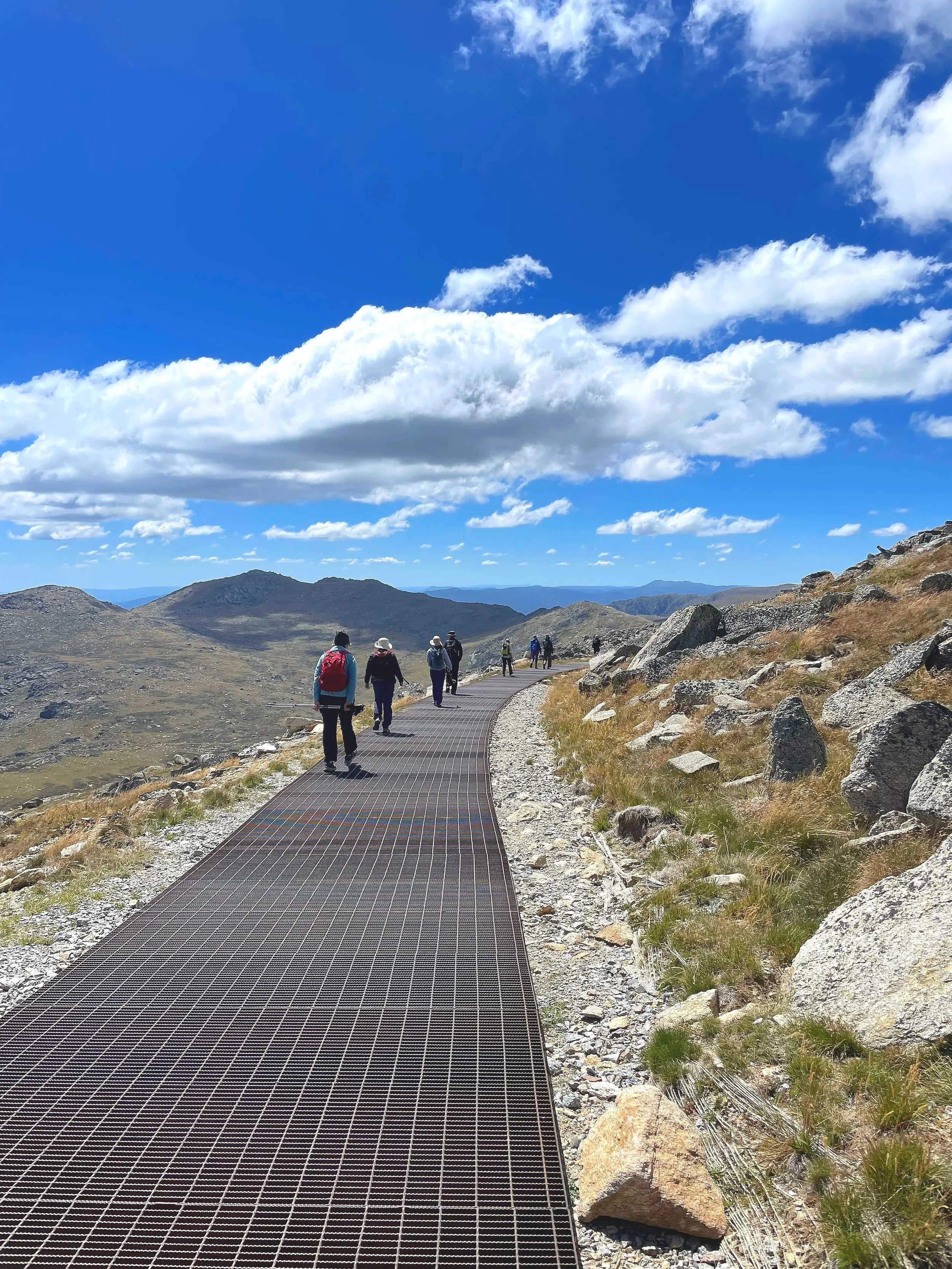 Hikers walking on a narrow metal trail in a mountainous, rocky landscape with a bright blue sky and scattered clouds overhead.