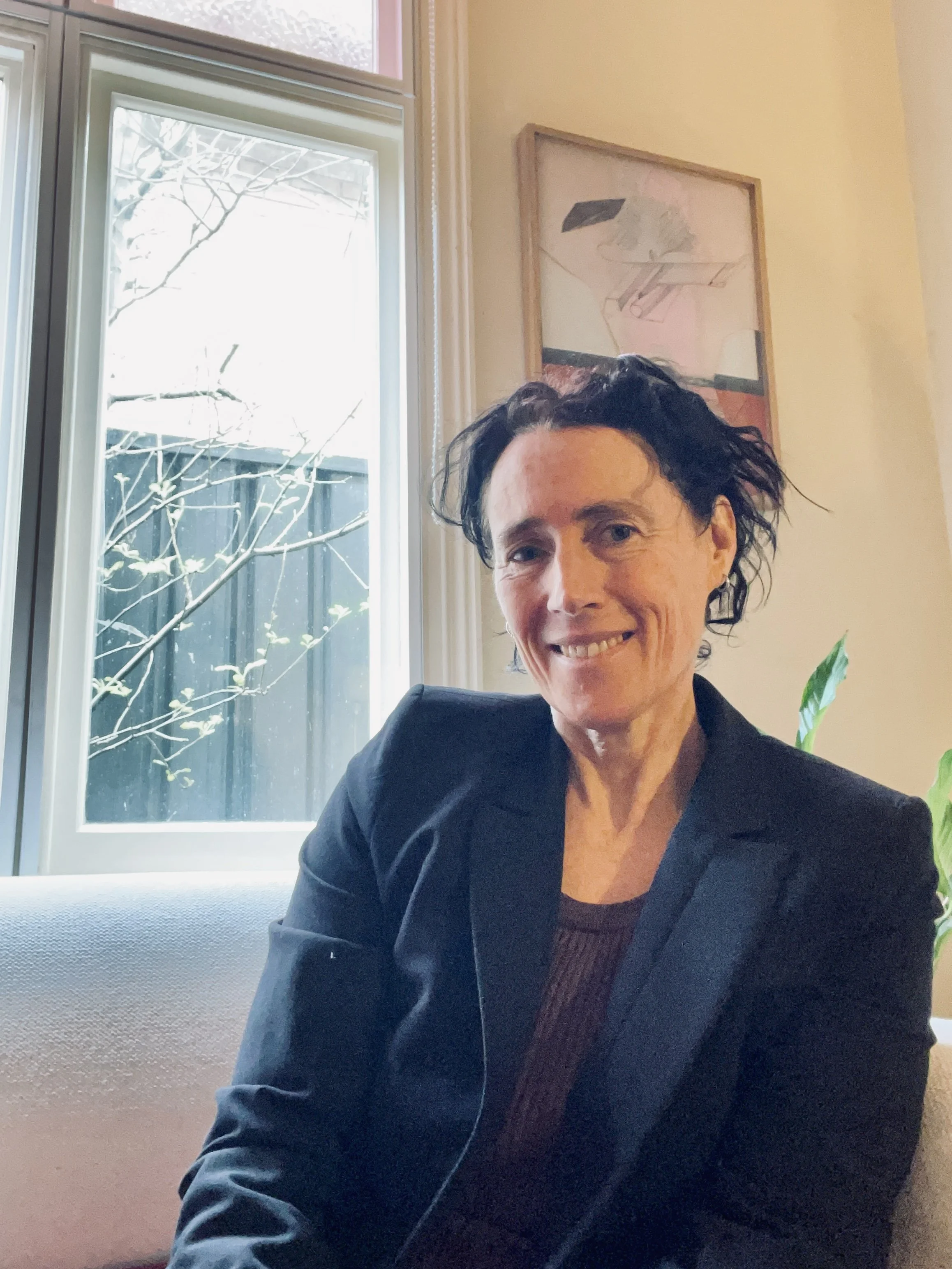 A woman with short, dark, curly hair smiling and wearing a black blazer sitting on a light-colored sofa near a window with a view of tree branches outside. There is a framed art piece on the wall behind her and a green plant next to her.