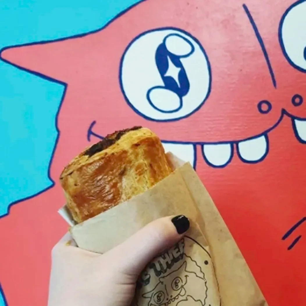 Hand holding a sausage breakfast sandwich in front of a colorful cartoon character with large eyes and teeth.