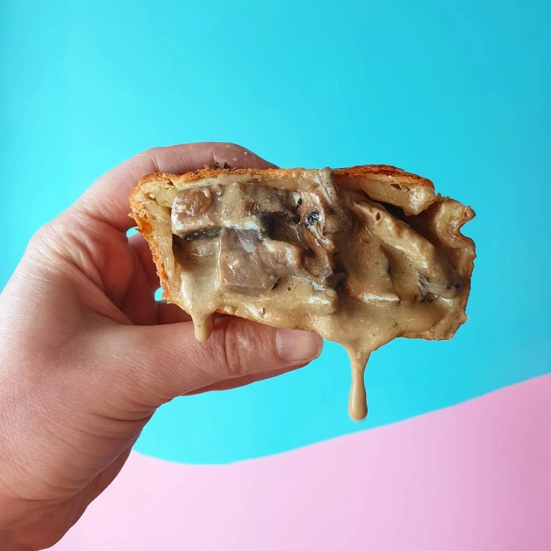 Slice of creamy mushroom pie held in hand against pastel blue and pink background.
