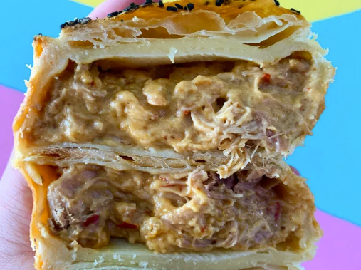 A hand holding a Thai red chicken pie with a golden, flaky crust topped with black sesame seeds, revealing the creamy chicken filling inside.