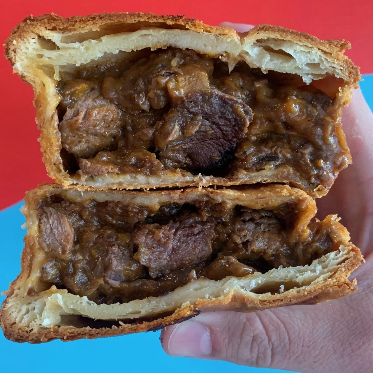 Close-up of a hand holding a sliced meat pie with a golden-brown crust and savory beef filling inside, against a colorful background.