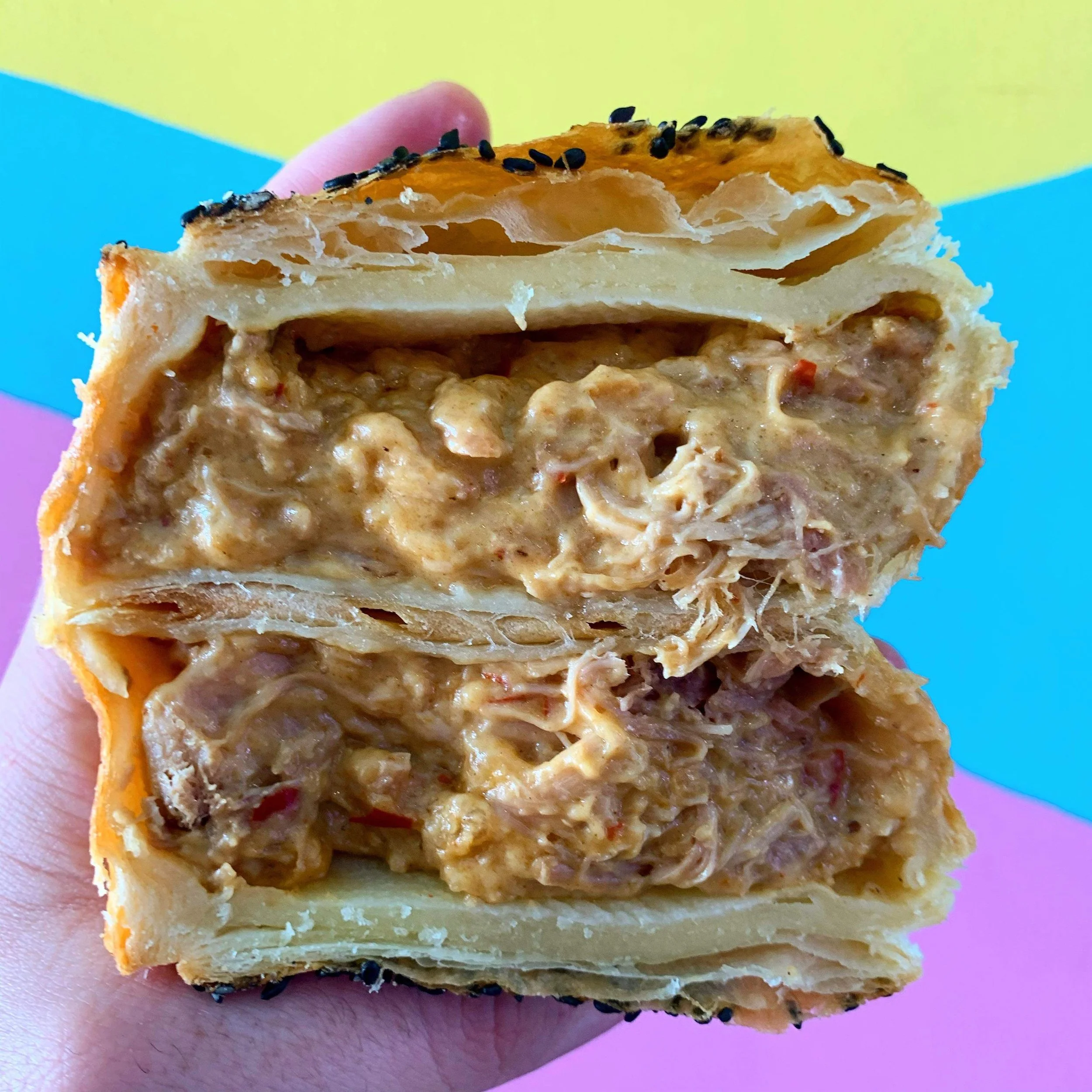 A hand holding a Thai red chicken pie with a golden, flaky crust topped with black sesame seeds, revealing the creamy chicken filling inside.