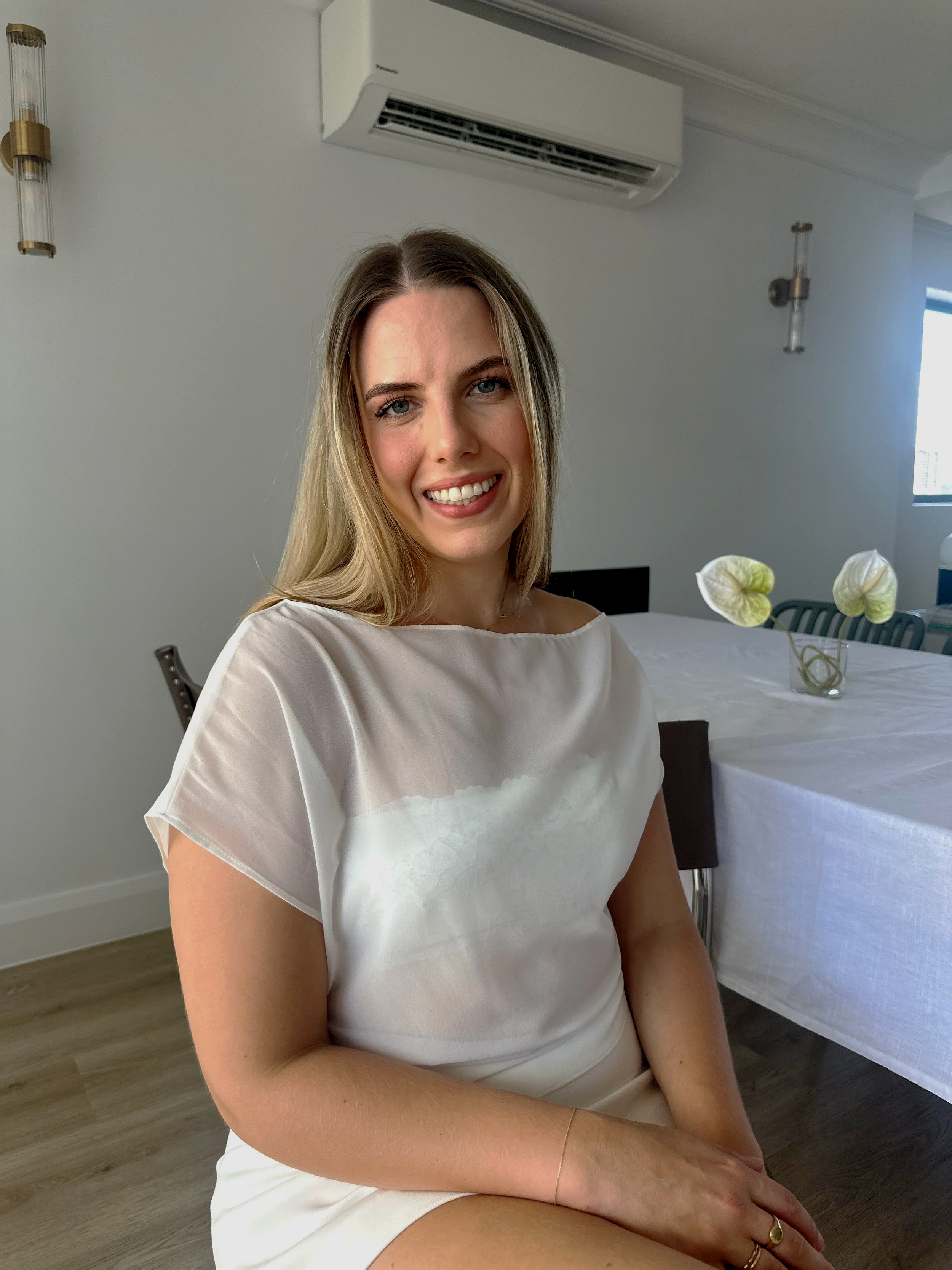 A woman with blonde hair, smiling, sitting in a dining room with white walls and a long white table with a glass vase and white flowers.