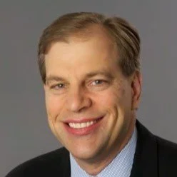 A smiling man with light brown hair wearing a dark blazer and light blue shirt against a gray background.
