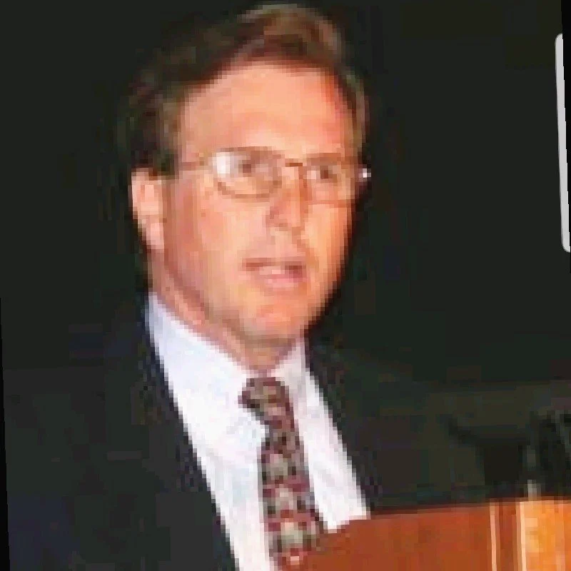 A man with glasses and brown hair wearing a suit and tie, speaking at a podium.