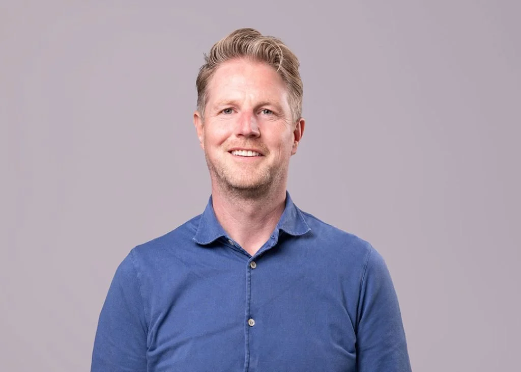 A smiling man with blonde hair in a blue button-down shirt, standing against a plain gray background.