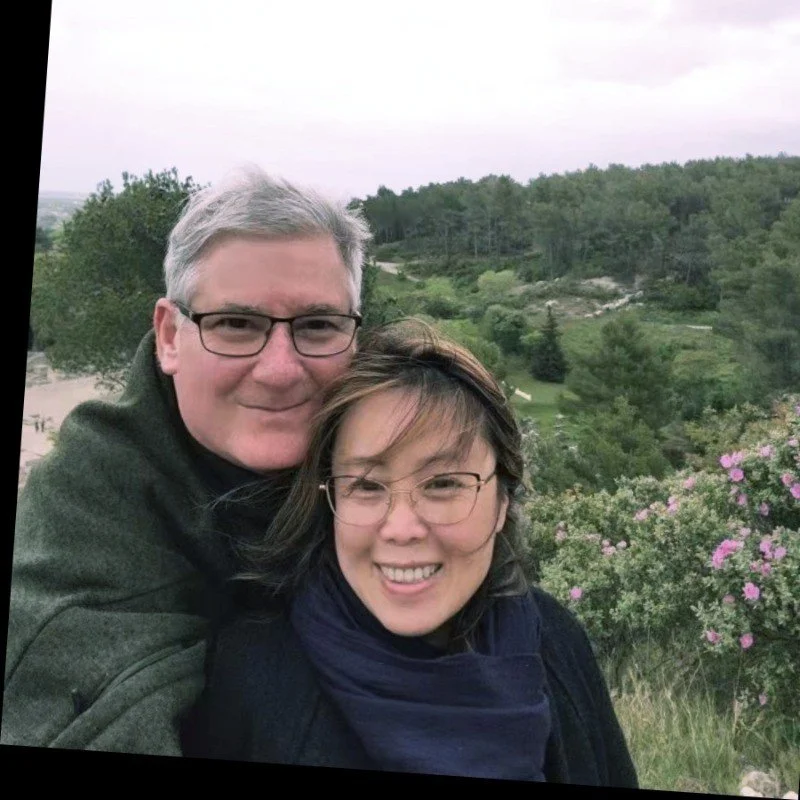 Two people taking a selfie outdoors with a scenic background of trees, greenery, and pink flowering bushes.