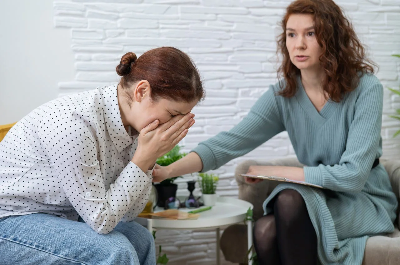 A woman is sitting on a sofa, comforting a young woman who is crying and covering her face with her hands. The comforting woman has a clipboard and is reaching out to the other woman.