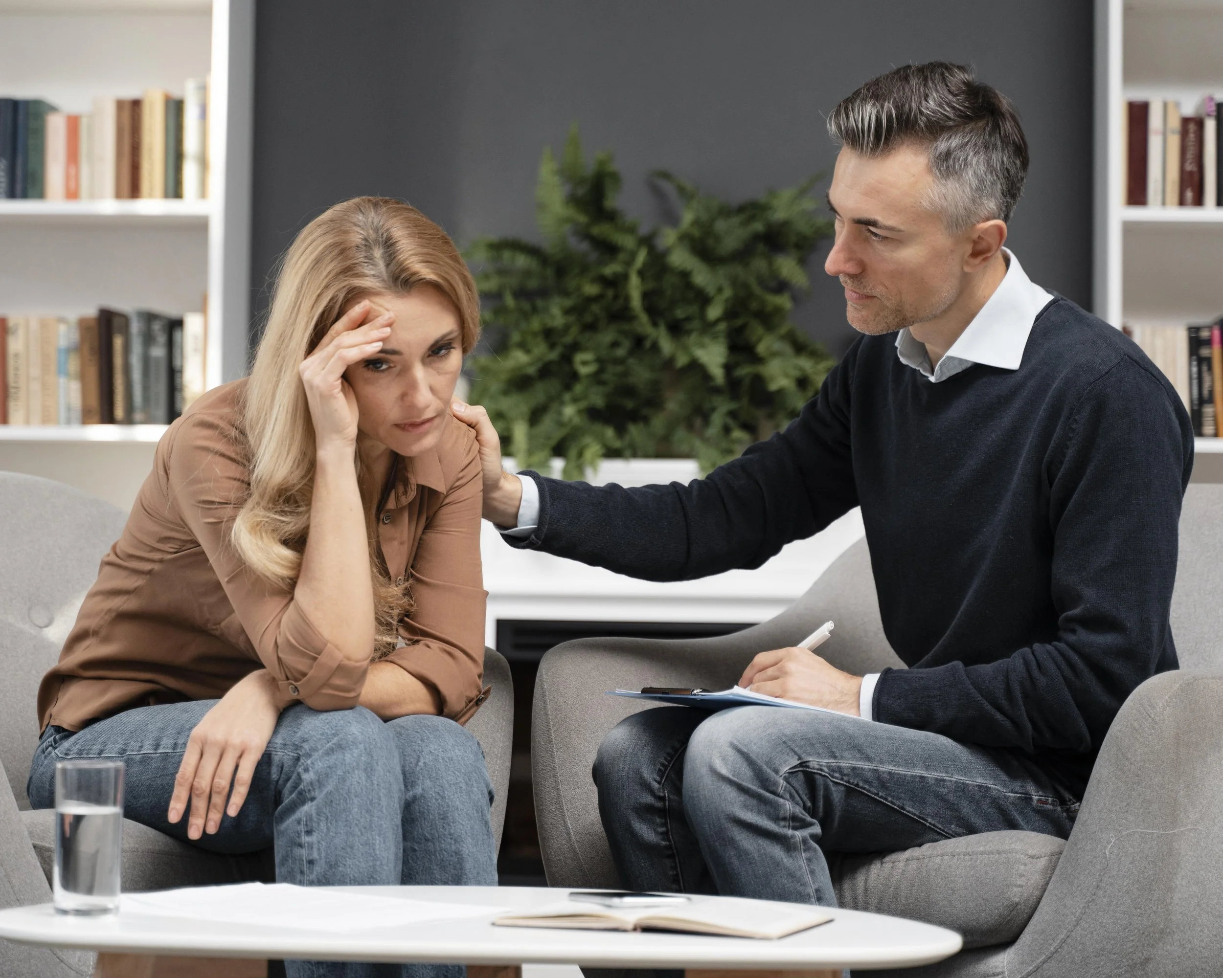 A woman sitting on a gray sofa with a distressed expression, holding her head, while a male counselor sitting in an armchair gently touches her shoulder in a therapy session in a room with bookshelves and a large plant in the background.