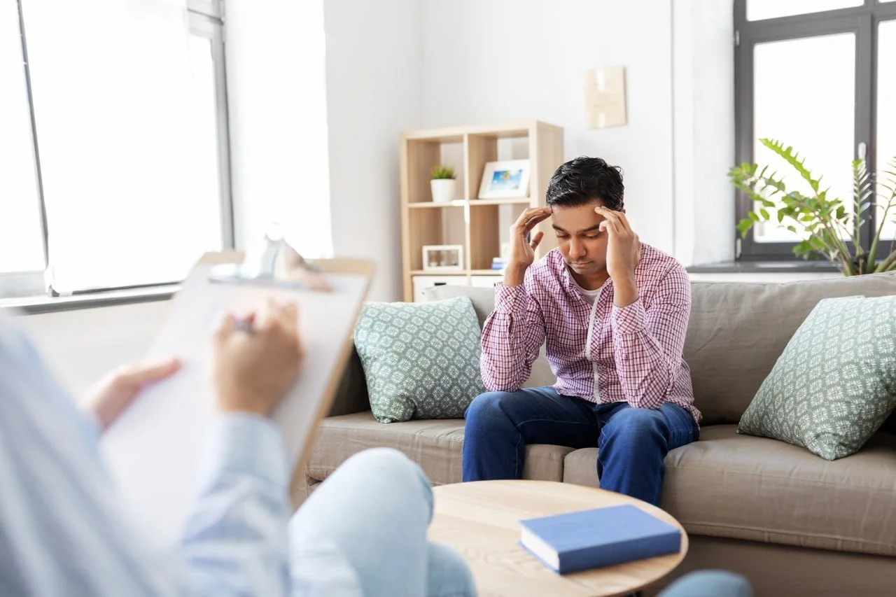 Man sitting on sofa during therapy session, pressing temples, with therapist taking notes, in a bright living room with large windows and houseplants.