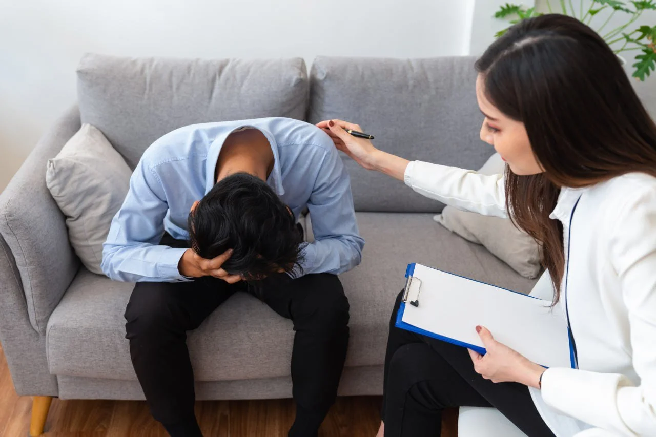 A woman in a white blazer sitting on a chair comforting a person seated on a gray sofa, who is hunched over with their head in their hand and appears distressed. The woman is holding a clipboard and has her hand on the person's shoulder, offering sup