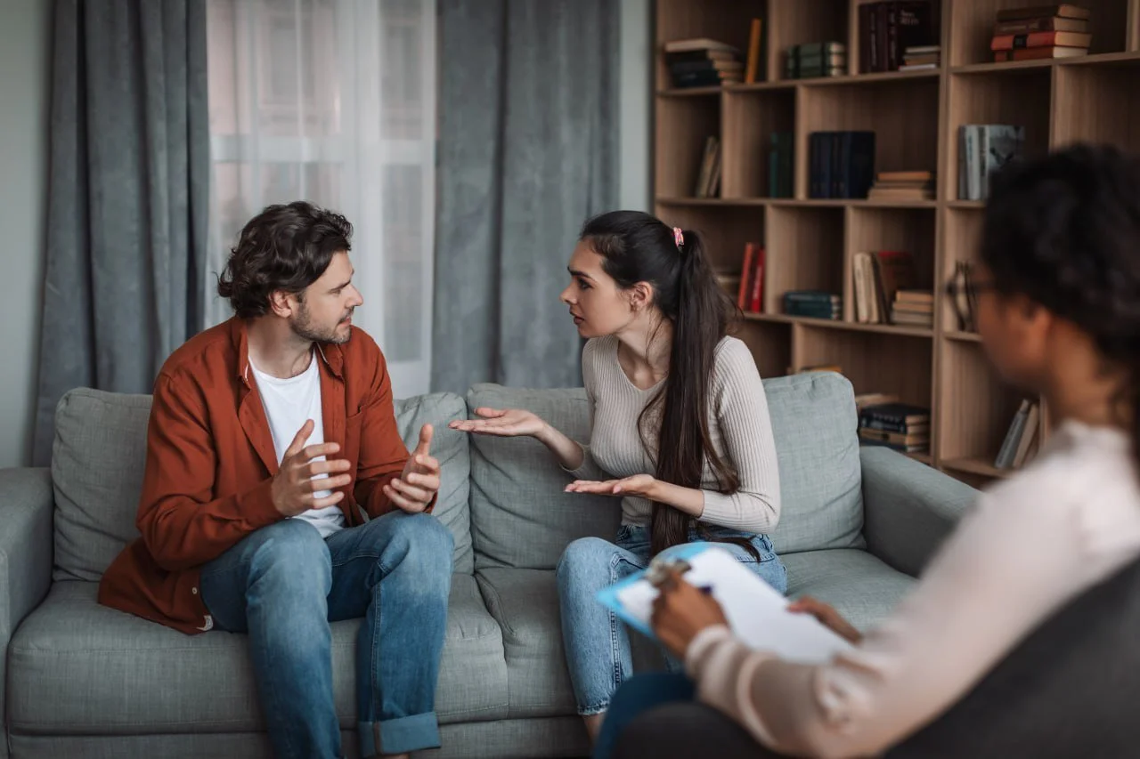 A woman and a man are having a serious discussion on a sofa, with a therapist taking notes during the session.