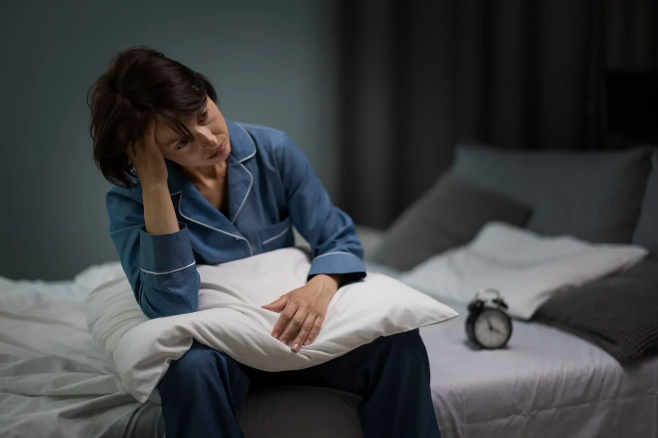 A woman in blue pajamas sitting on a bed holding her head and looking distressed, with a pillow on her lap. An alarm clock and pillows are on the bed behind her.