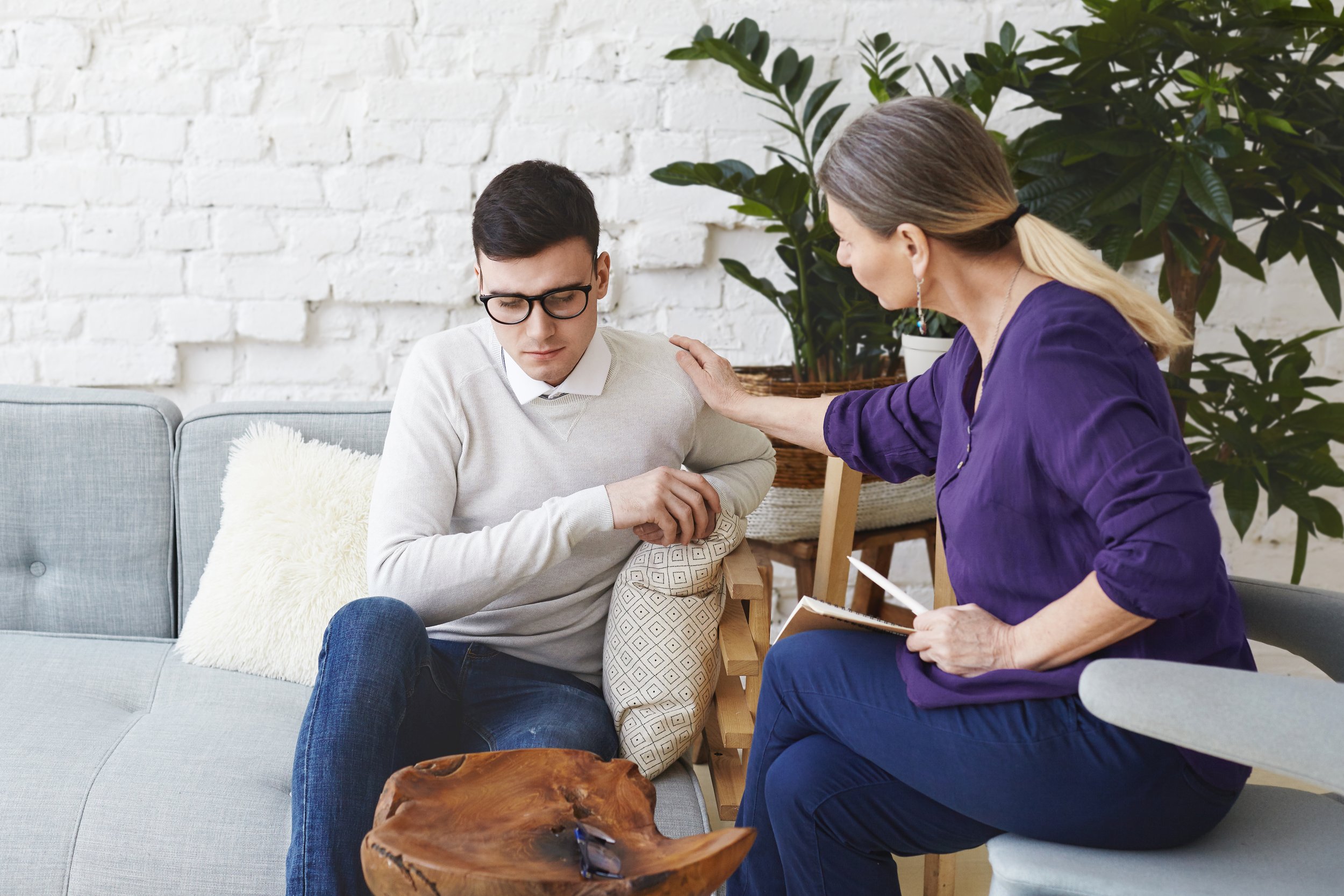 A young man with glasses and a white sweater sitting on a sofa with a hand on his shoulder, appearing distressed, as a woman in a purple blouse and blue pants offers comfort with a hand on his shoulder and holds a notebook and pen in her lap. They ar
