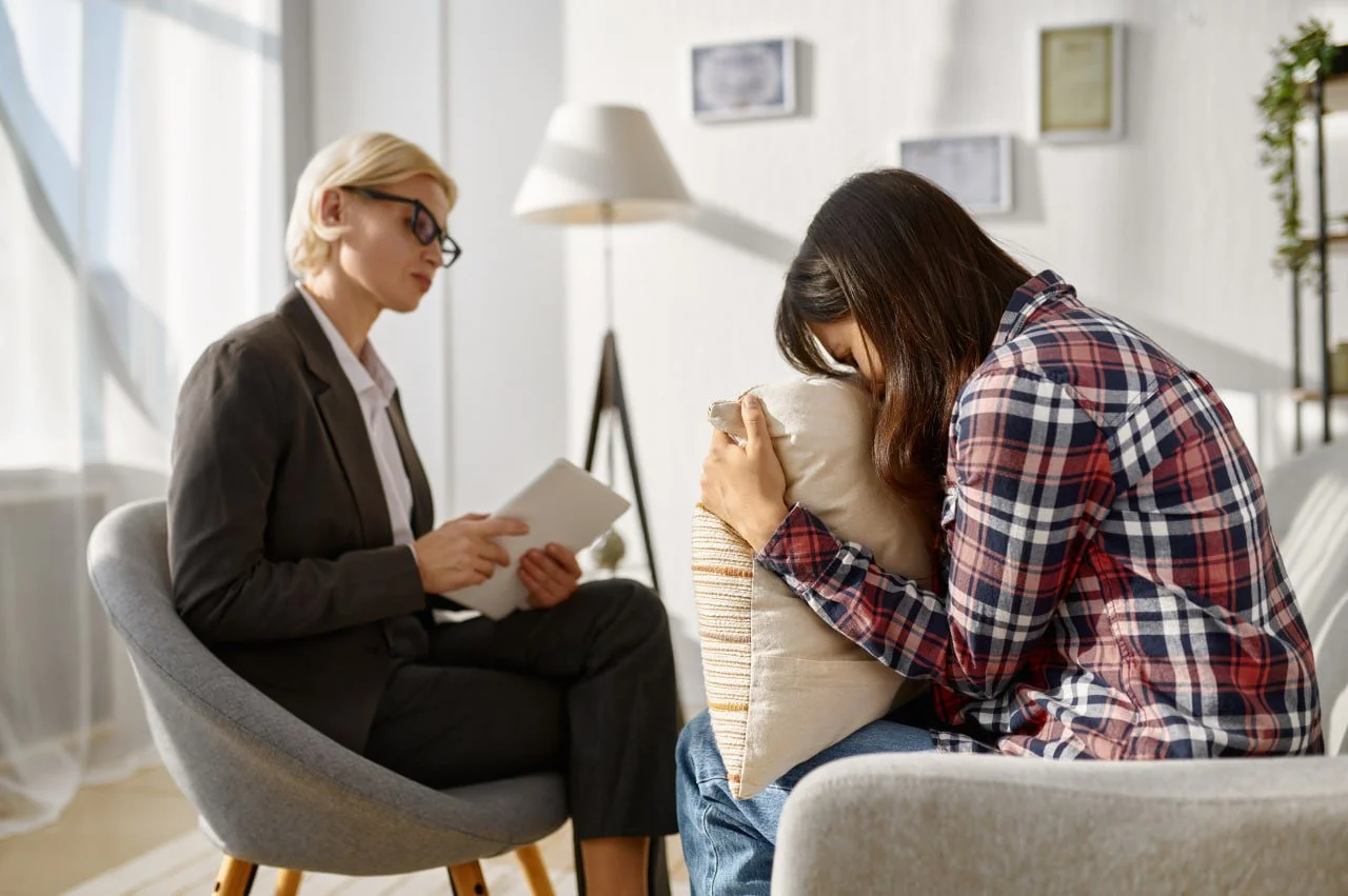 A woman in a plaid shirt is upset and crying into a pillow during a therapy session with a therapist, a woman in a business suit, in a cozy, well-lit room.