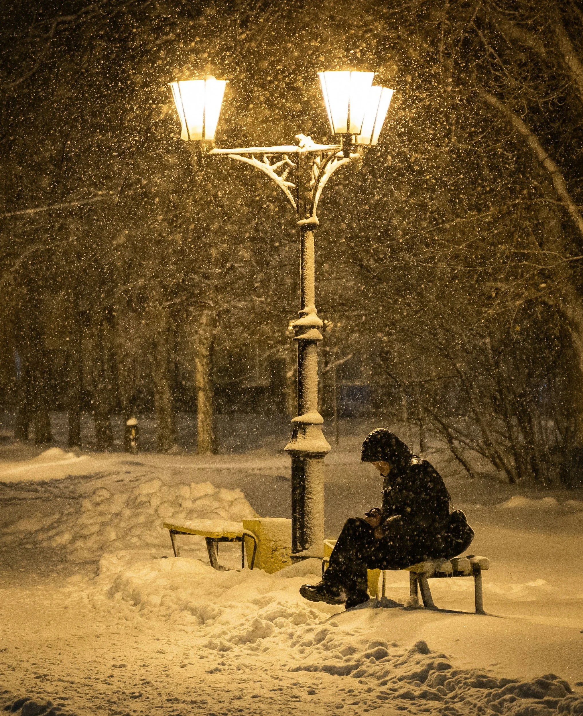 Individual sitting alone on a snow-covered bench at night during a snowfall, symbolizing winter isolation, seasonal loneliness and emotional hardship.