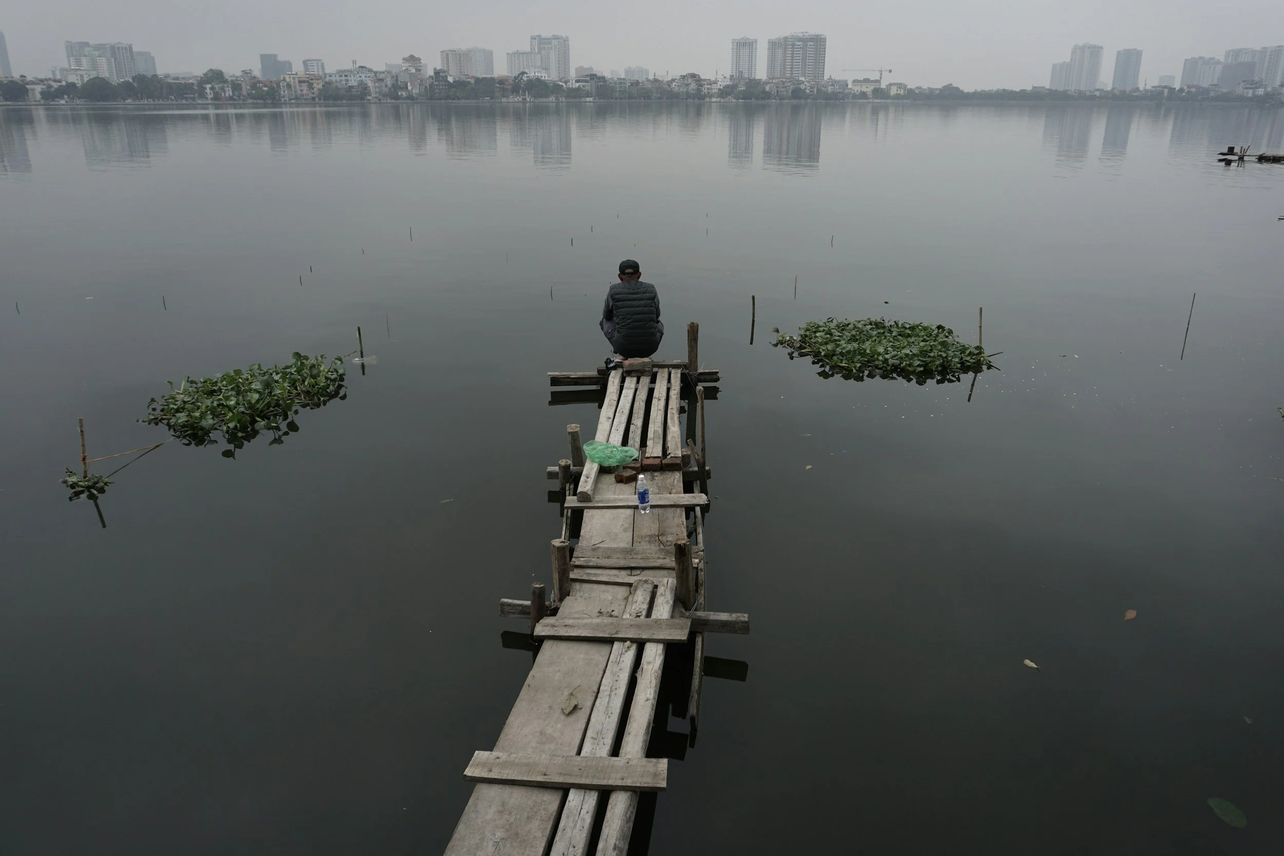 Person sitting alone on a wooden dock overlooking still water and distant city skyline, representing emotional isolation and reflective solitude.