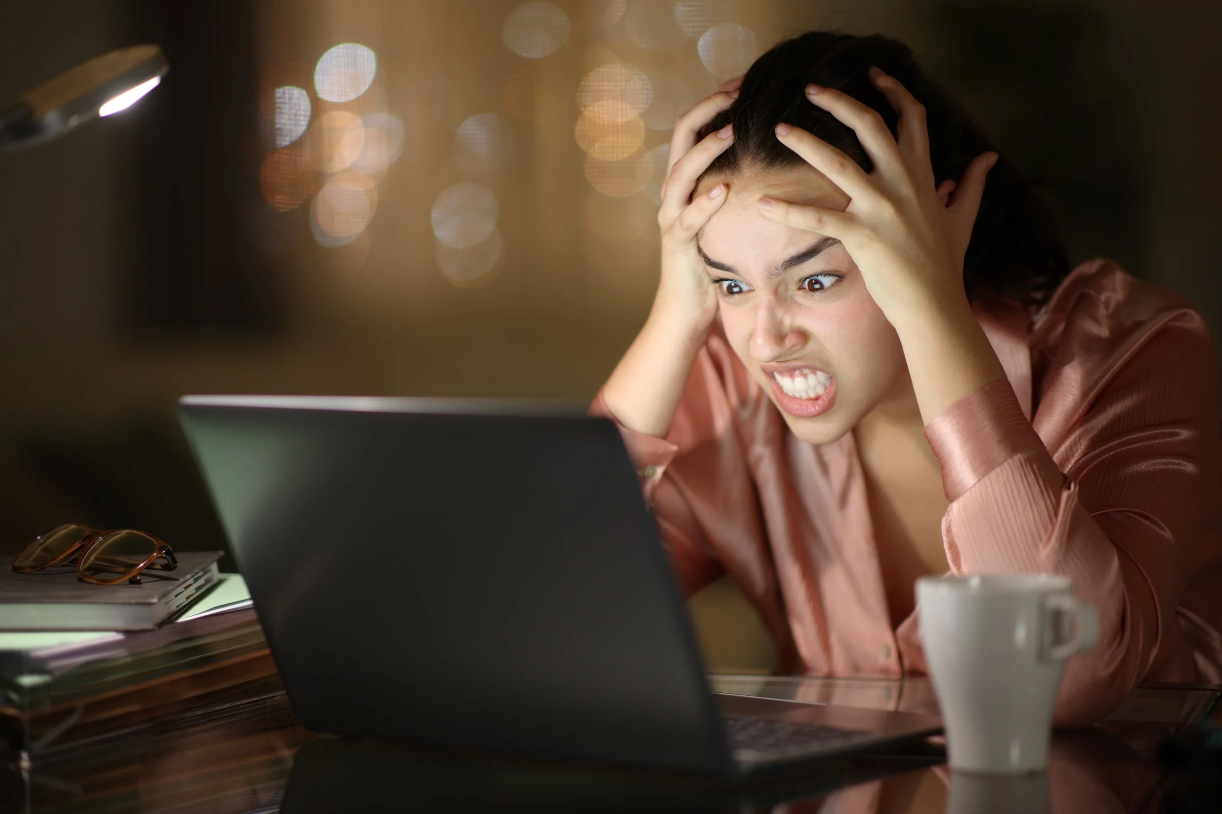 Frustrated woman with hands on her head looking at a laptop in a dimly lit room