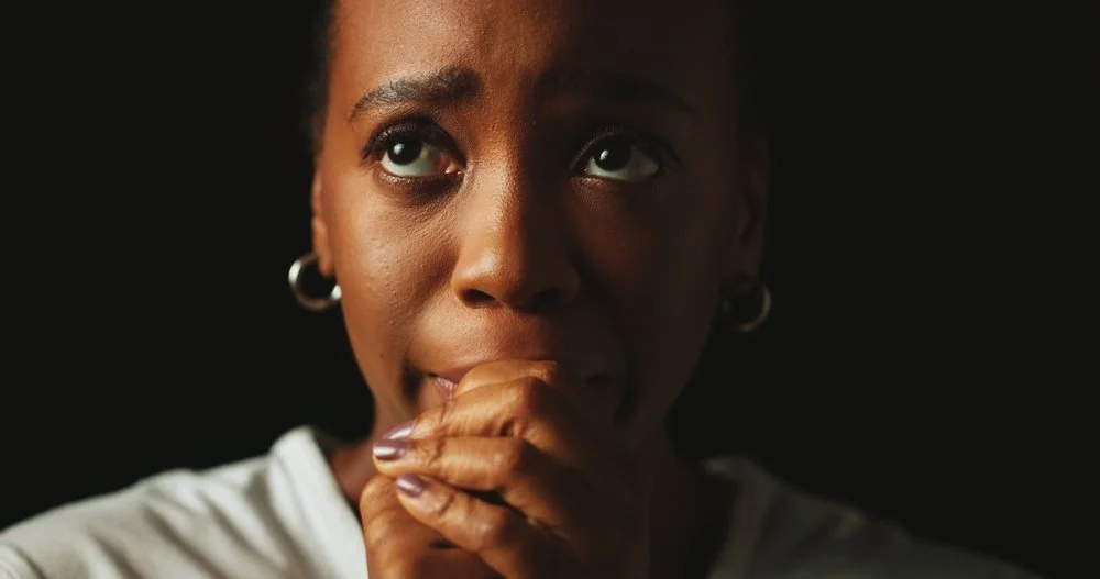 Close-up of a woman with short hair, wearing hoop earrings, resting her chin on her hand, looking thoughtful against a dark background.