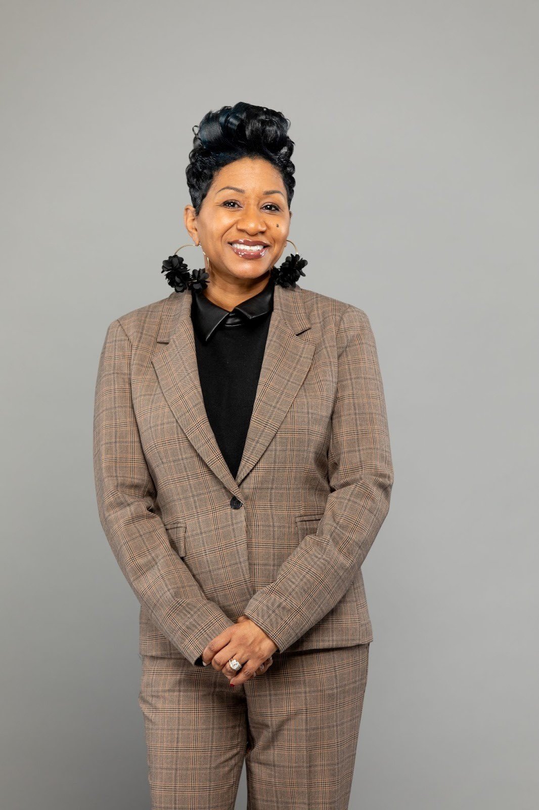 A smiling woman with short curly hair, wearing a brown plaid pantsuit and a black shirt with a collar, standing against a plain gray background.