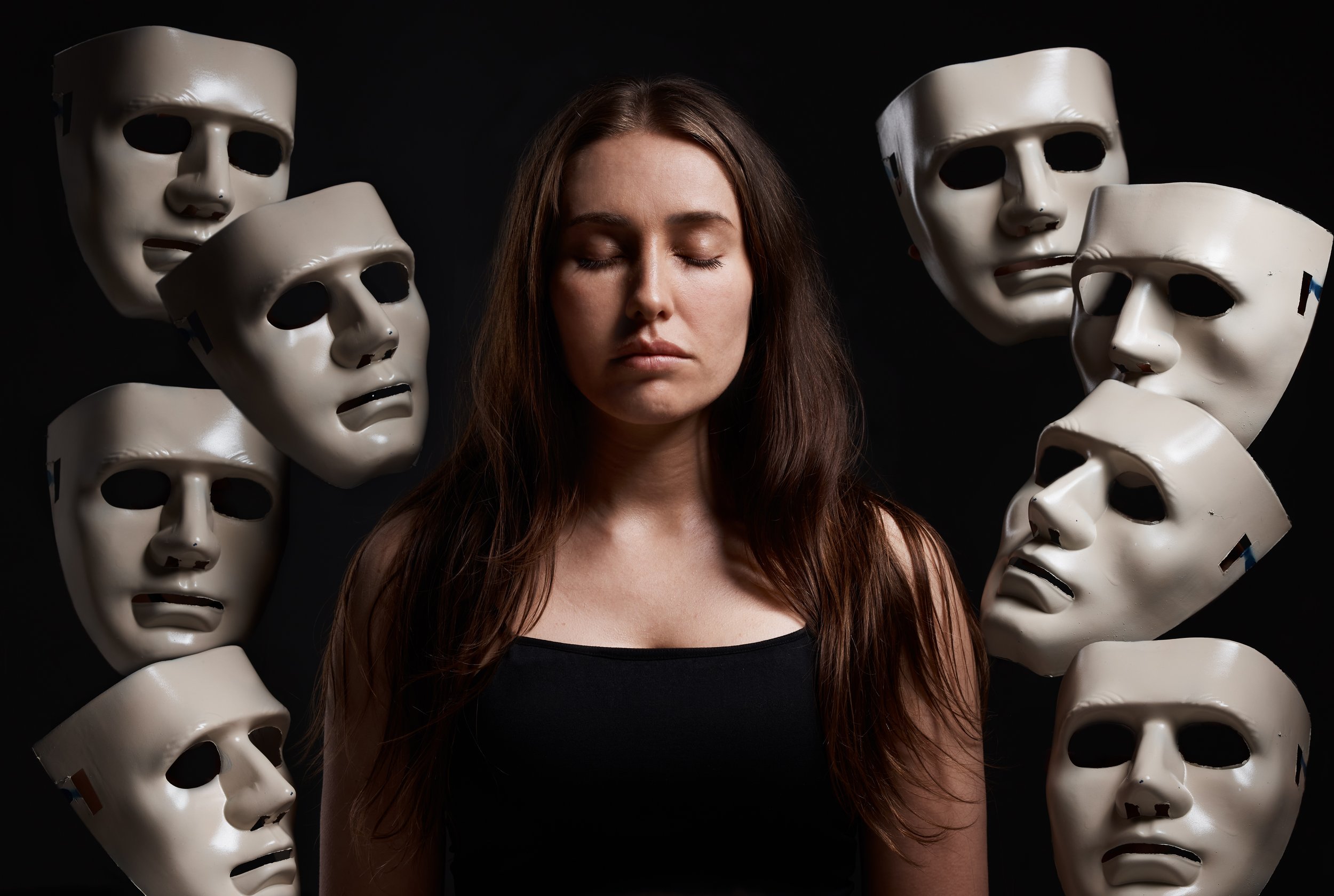 A woman with closed eyes and long brown hair stands in front of a black background, surrounded by white theatrical masks with various facial expressions.