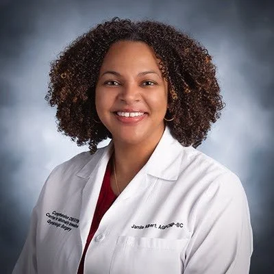 A woman with curly hair wearing a white medical coat smiling against a gray background.