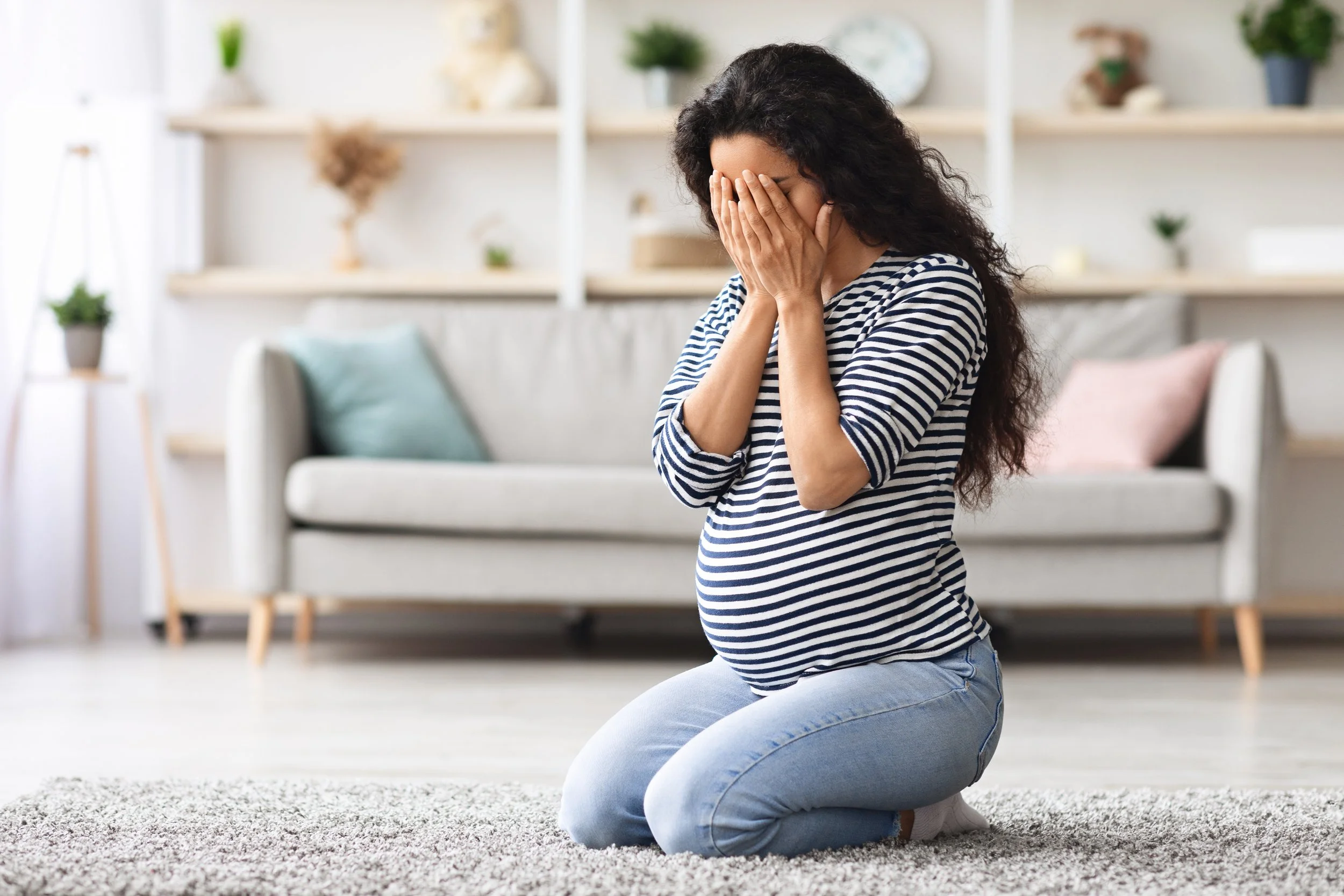 Pregnant woman kneeling on the floor, holding her face in her hands in a living room.