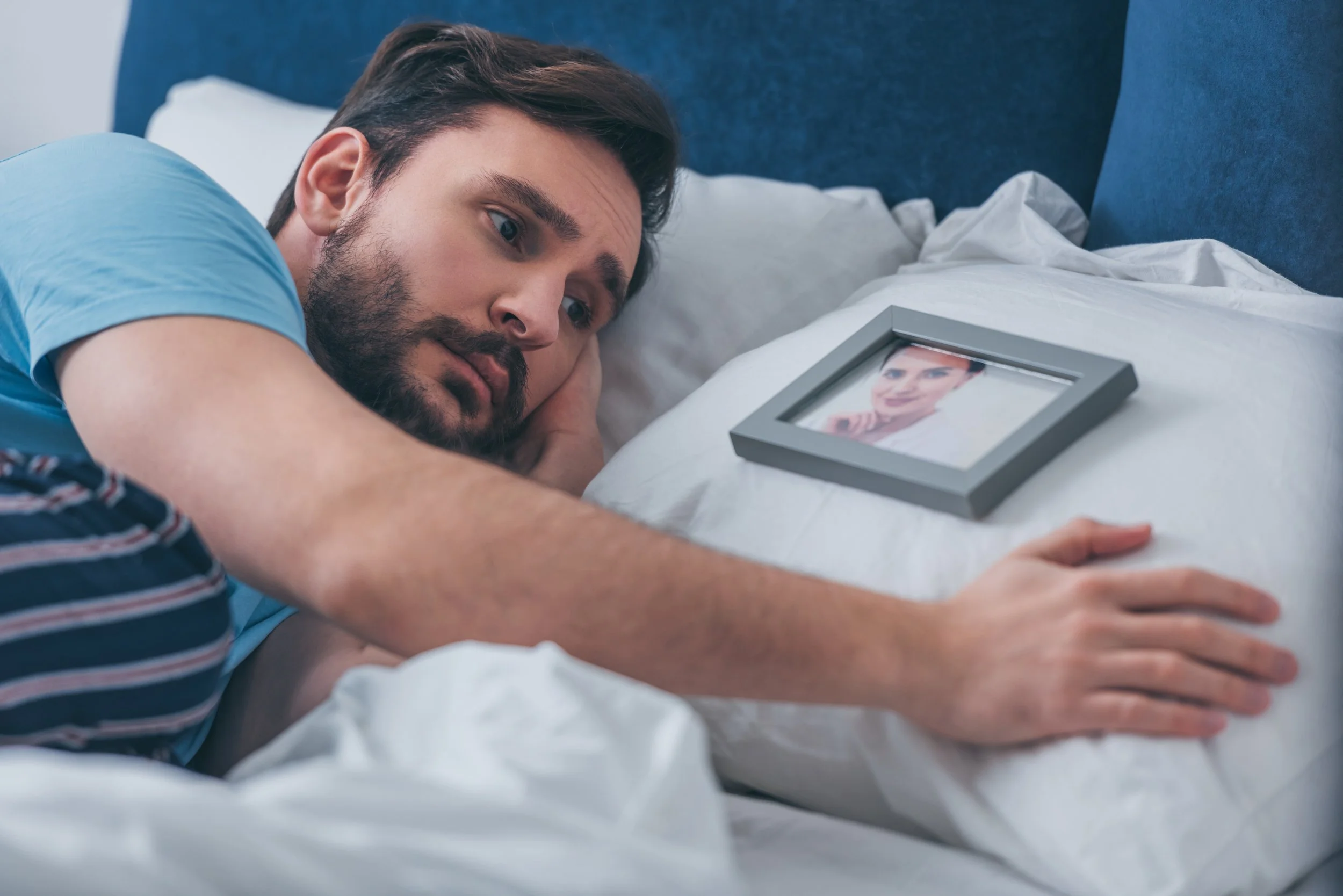 A man with dark hair and a beard lying on a bed, resting his head on a pillow, looking at a photograph of a woman in a gray frame on the bed.