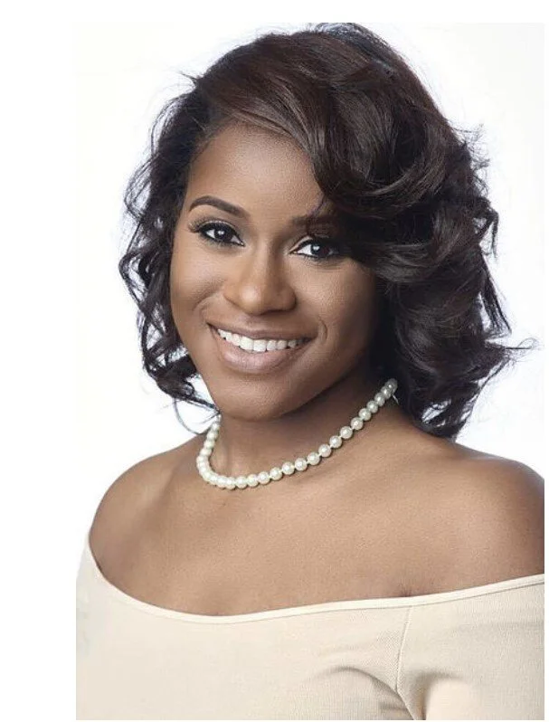 Portrait of a smiling woman with dark, curly hair wearing a pearl necklace and an off-the-shoulder white top against a white background.
